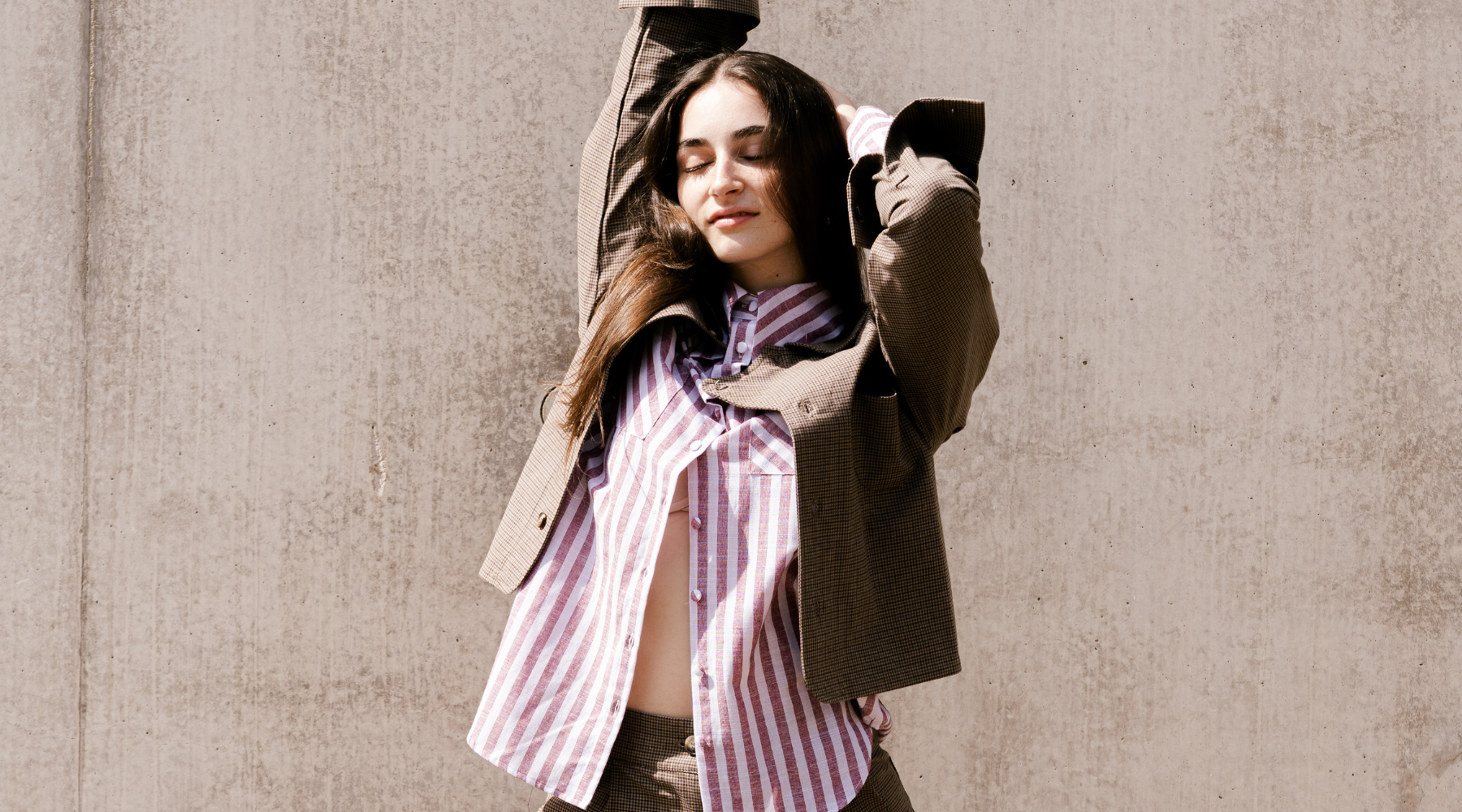 a woman wearing a stripe red shirt and a brown jacket, her eyes closed and the sun on her face, standing against a concrete wall