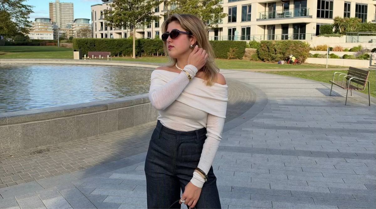 A woman stands outdoors near a paved walkway and water feature, wearing an off-the-shoulder cream long-sleeve top, dark wide-leg jeans, black sunglasses, and holding a small black handbag.