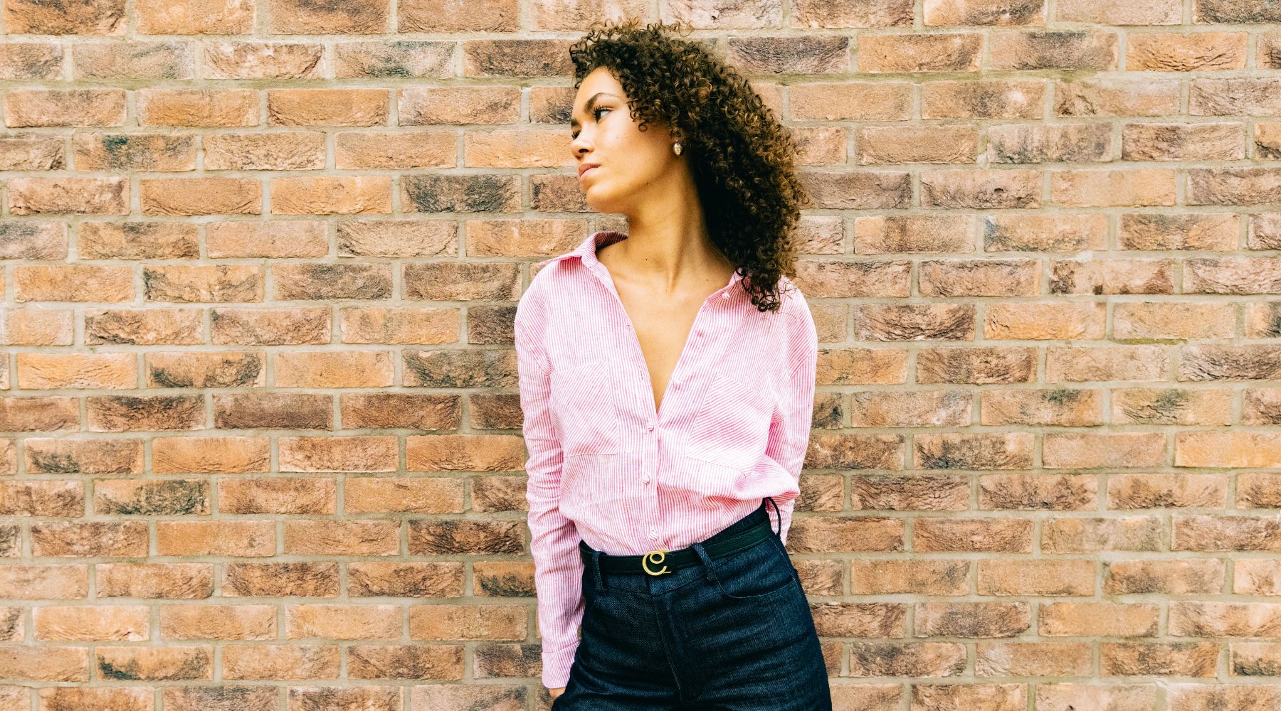 woman with black curly hair wearing pink shirt, dark trouser and brown belt against a brick wall
