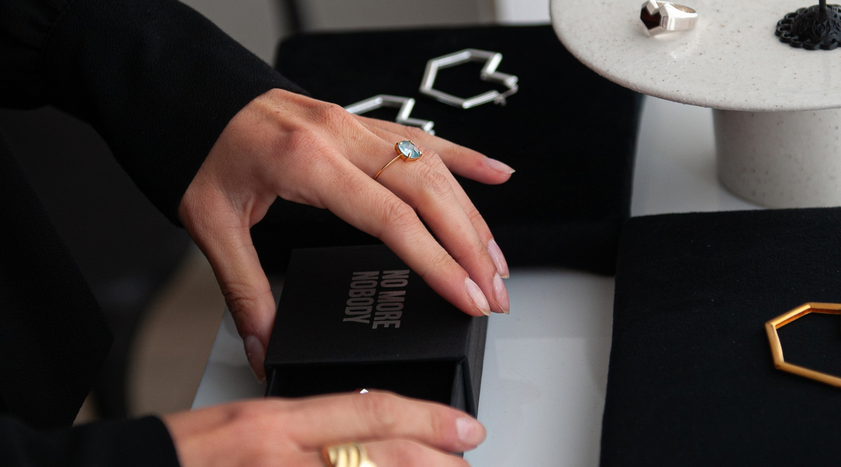 jewelry display with silver and gold jewelry and woman hands opening a gift box with No MORE NOBODY brand on it.