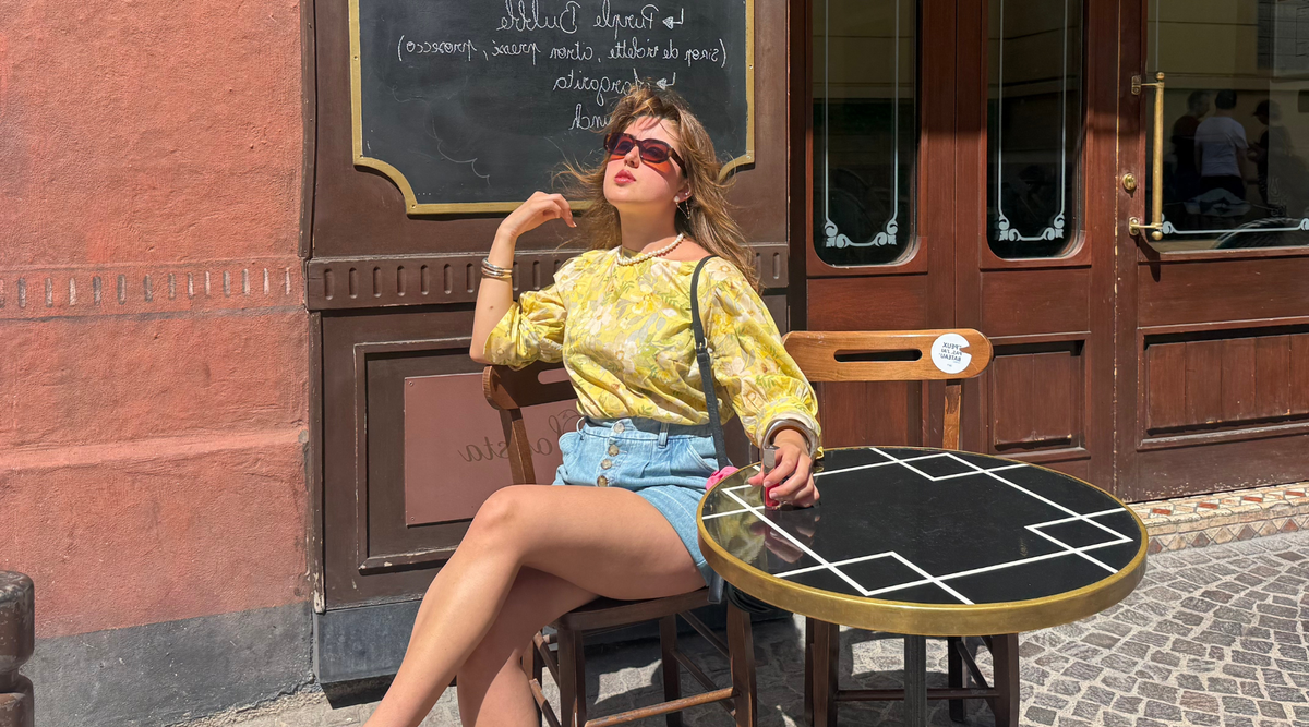 woman moving her hair away from her face sitting outside a restaurant wearing a yellow floral top and denim shorts