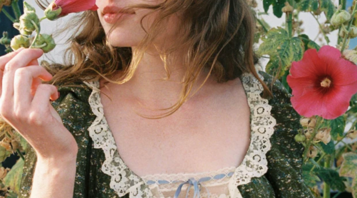 woman holding a green small plant with red flowers around her, wearing a green open neck dress with frills around