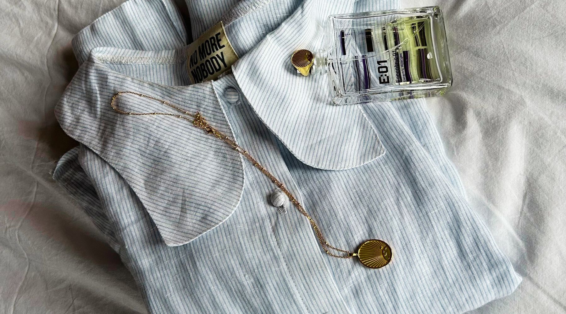 blue stripe shirt folded on a white linen bed, with perfume bottle, gold necklace and gold ring