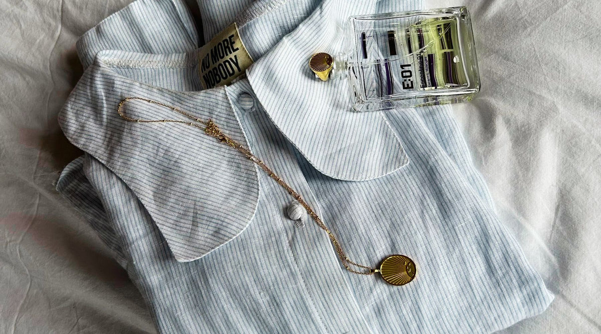 blue stripe shirt folded on a white linen bed, with perfume bottle, gold necklace and gold ring