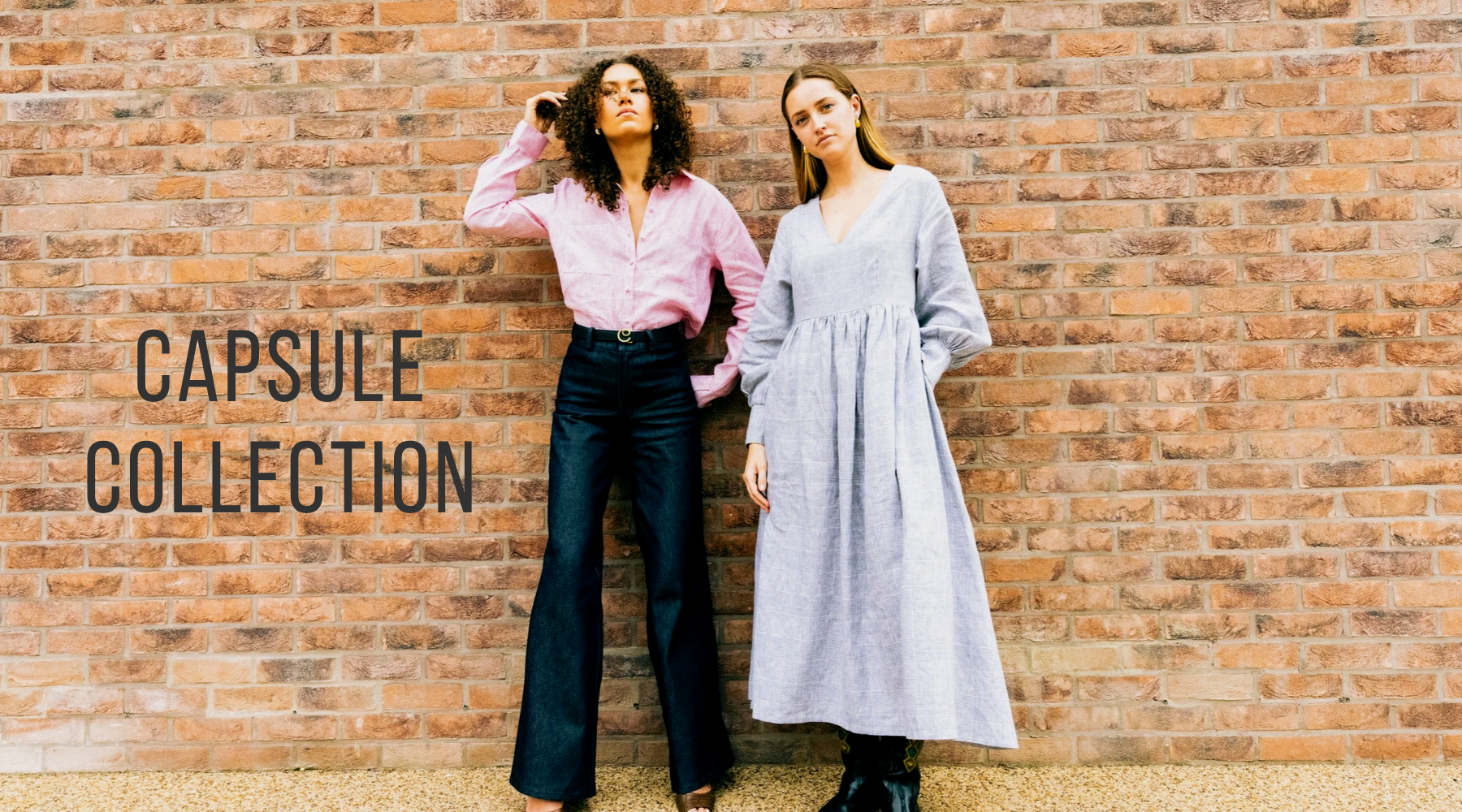 two women wearing dark jeans and red shirt, the other wearing a grey maxi dress against a brick wall