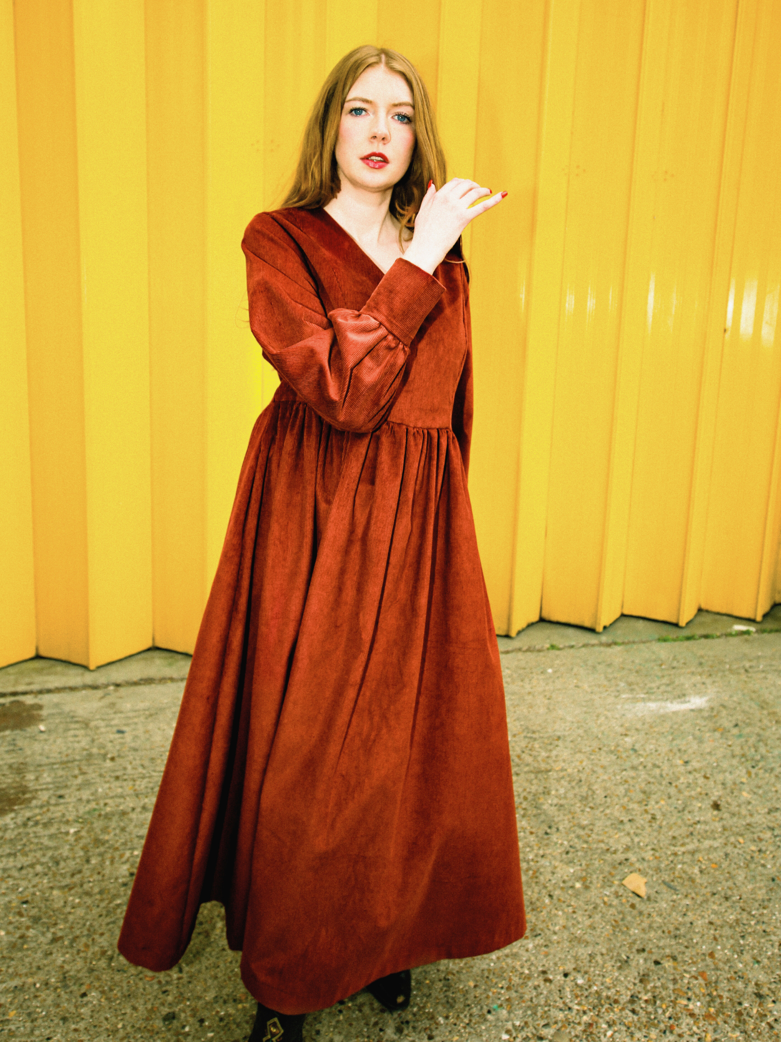 Woman wearing a long rust red dress against a yellow wall.
