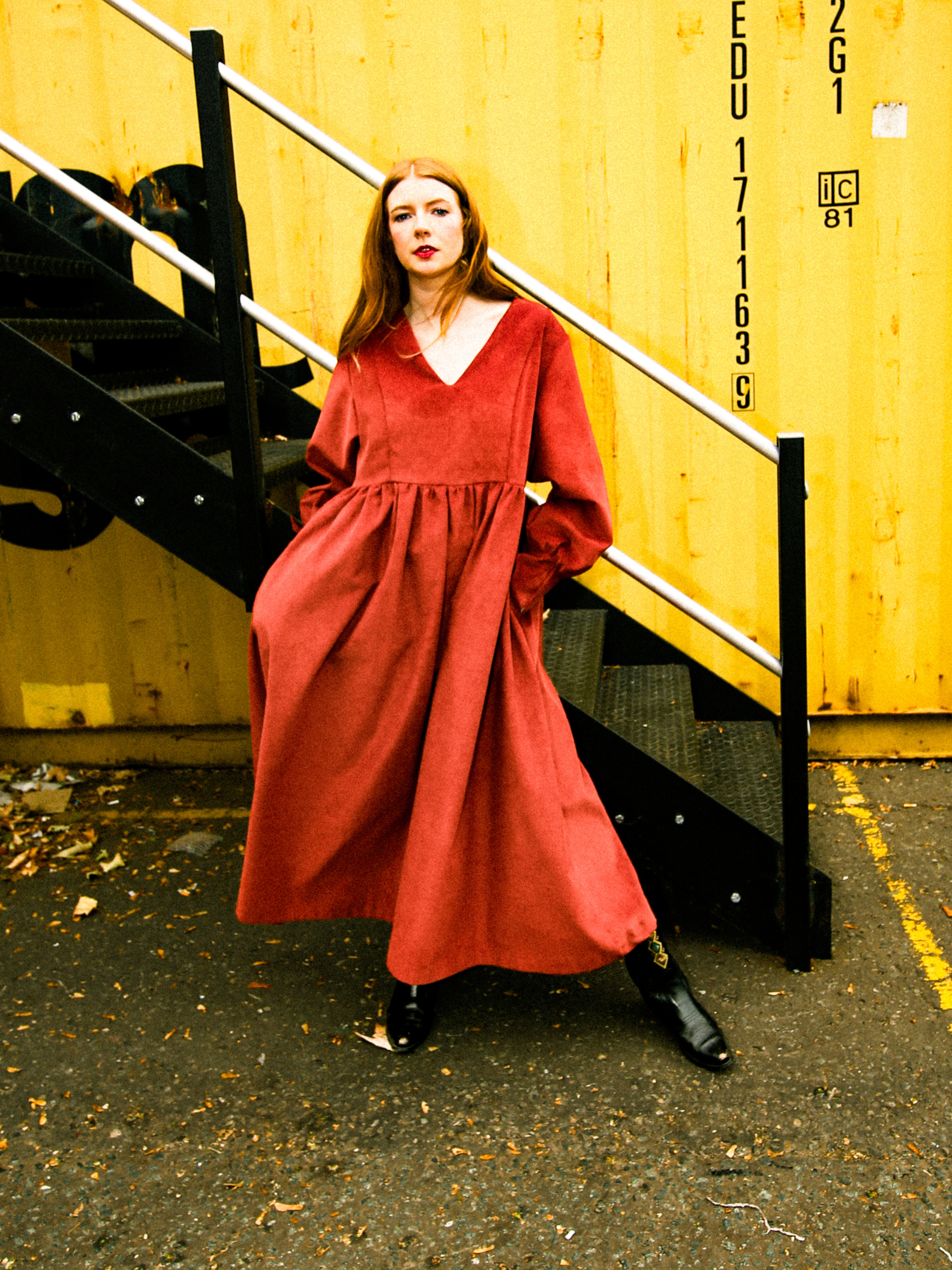 Woman in a red rust dress standing on a black staircase against a yellow wall with text.