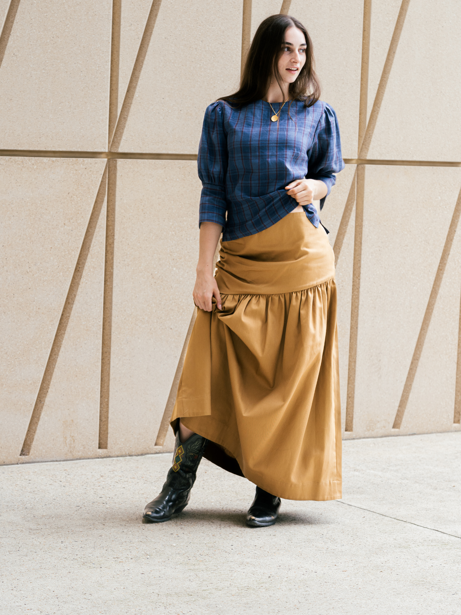 Woman wearing a blue plaid shirt and tan skirt against a geometric wall.