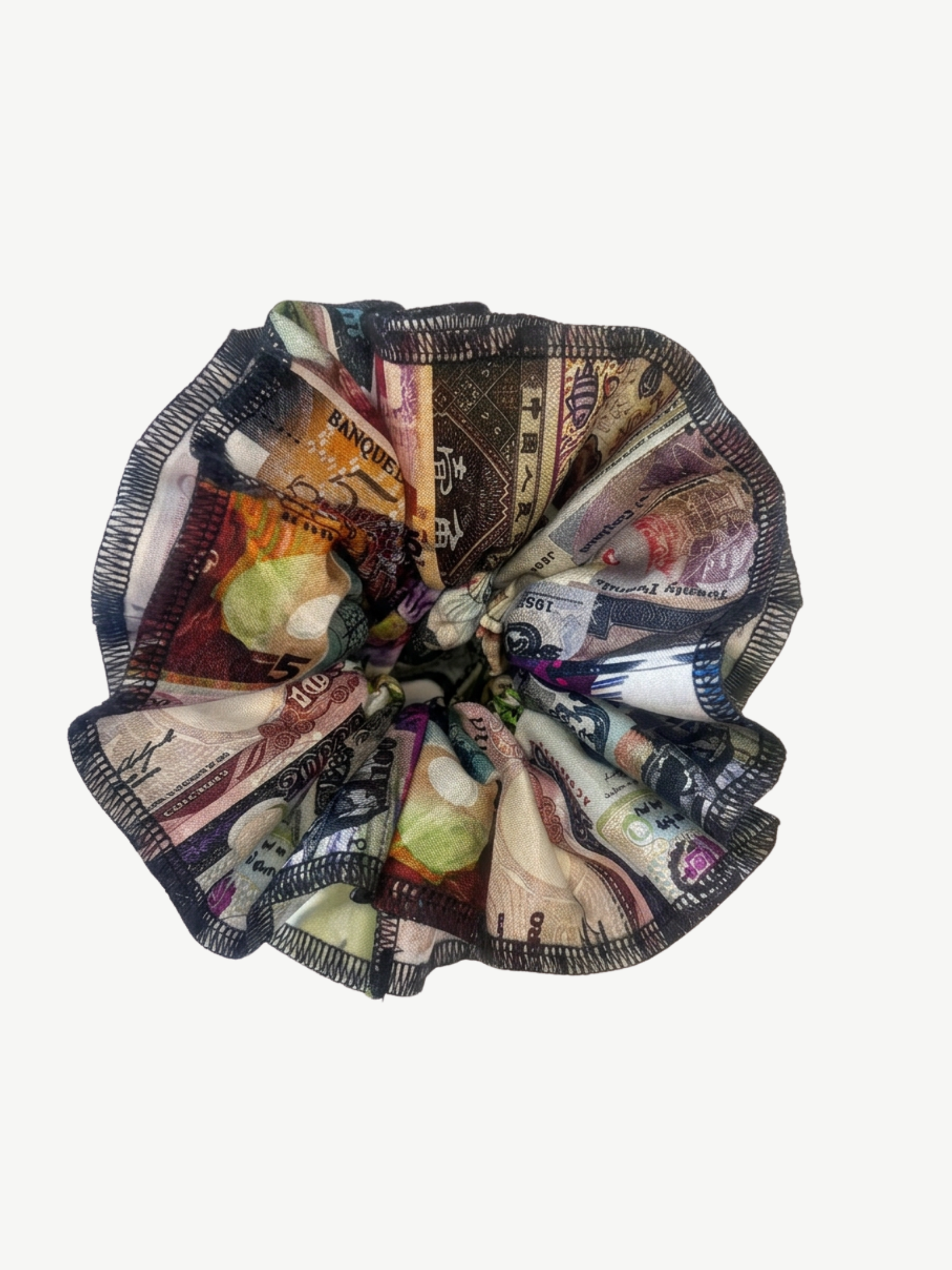 A fabric hair scrunchie featuring an all-over print of various banknotes and currency notes, finished with a black and white striped braided trim, shown on a plain light gray background.