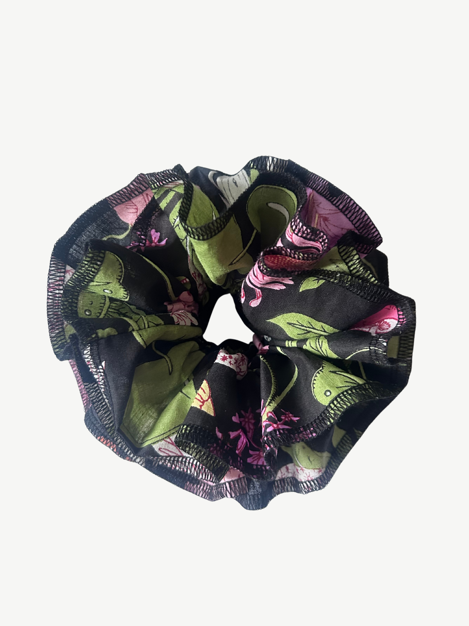 A fabric hair scrunchie featuring a botanical print with green leaves, pink flowers, and black mesh-like detailing, shown against a plain light gray background.