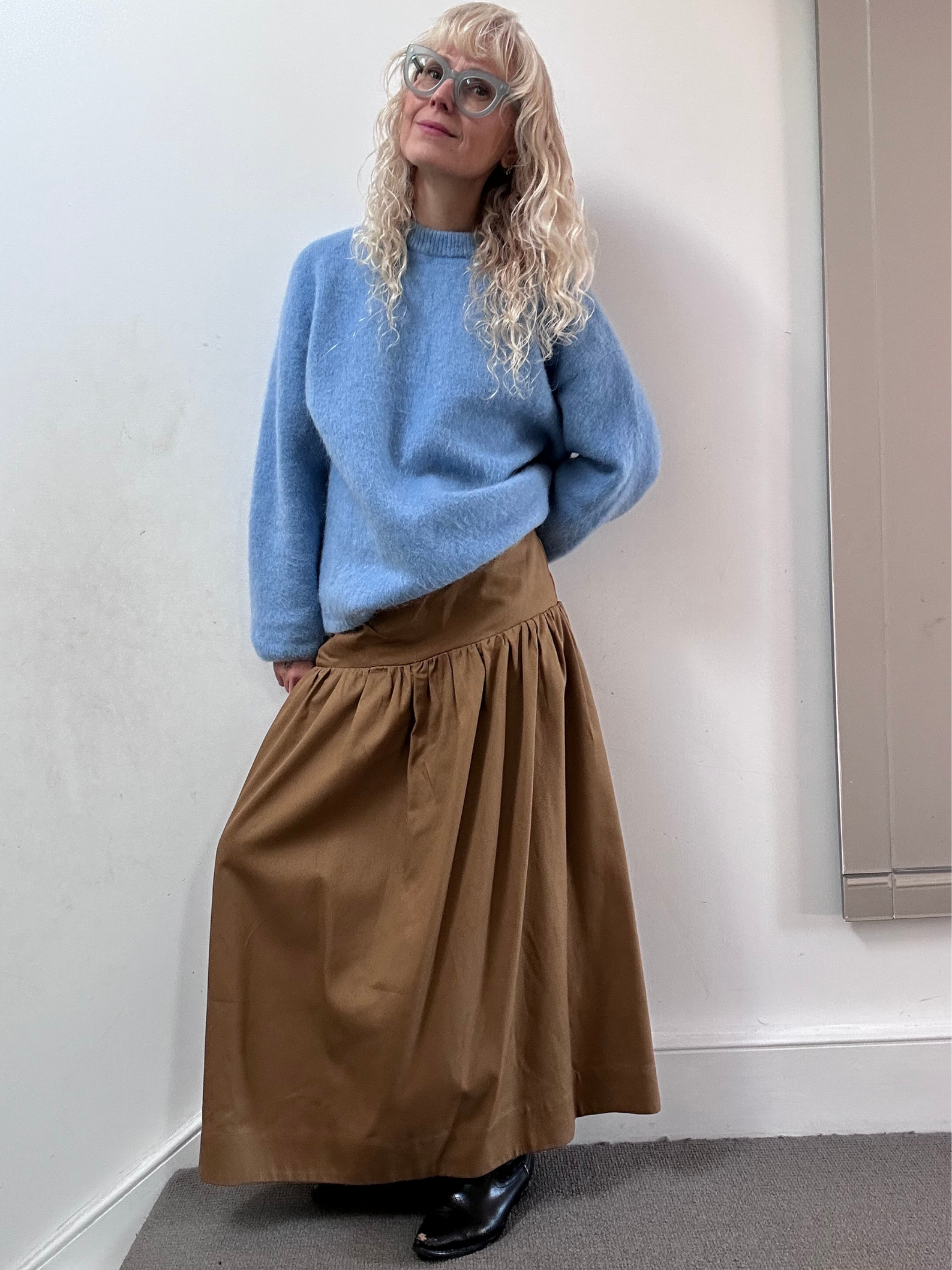 Person wearing a blue sweater and brown skirt against a white wall.