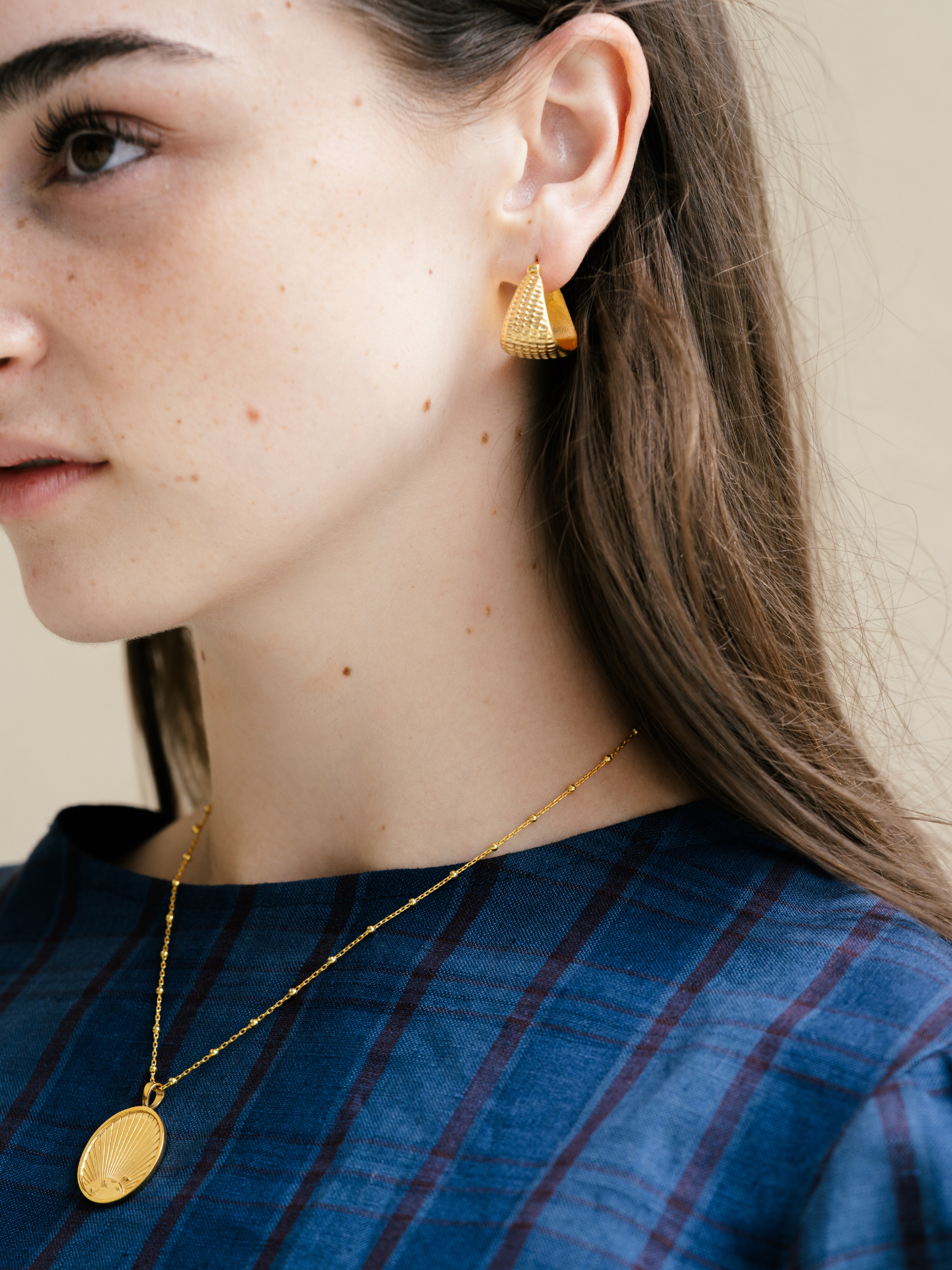 Close-up of a person wearing gold earrings and a necklace against a neutral background
