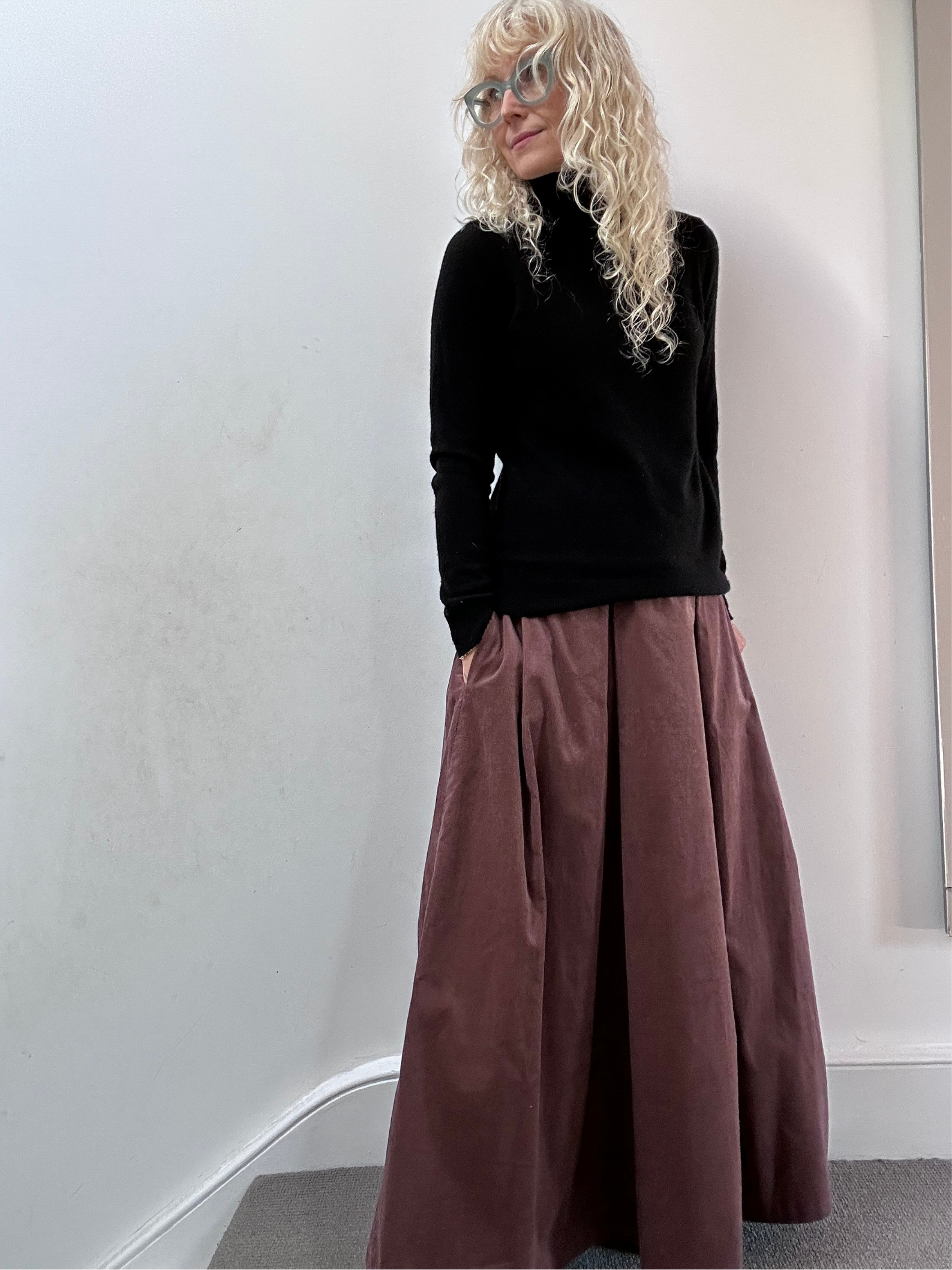 Person wearing a black top and long pink skirt against a white wall.