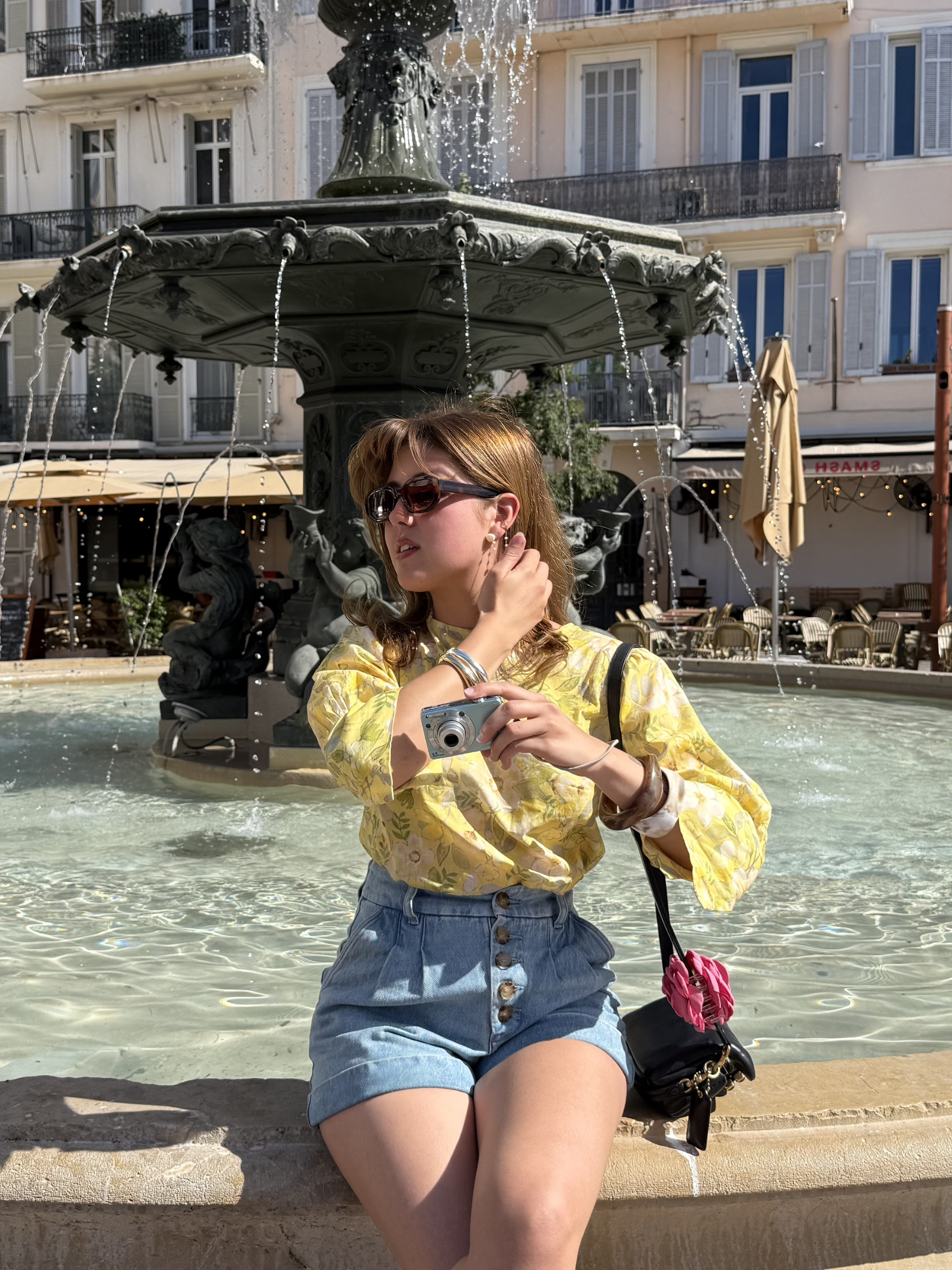 Woman in yellow blouse and denim shorts taking a photo in front of a fountain.