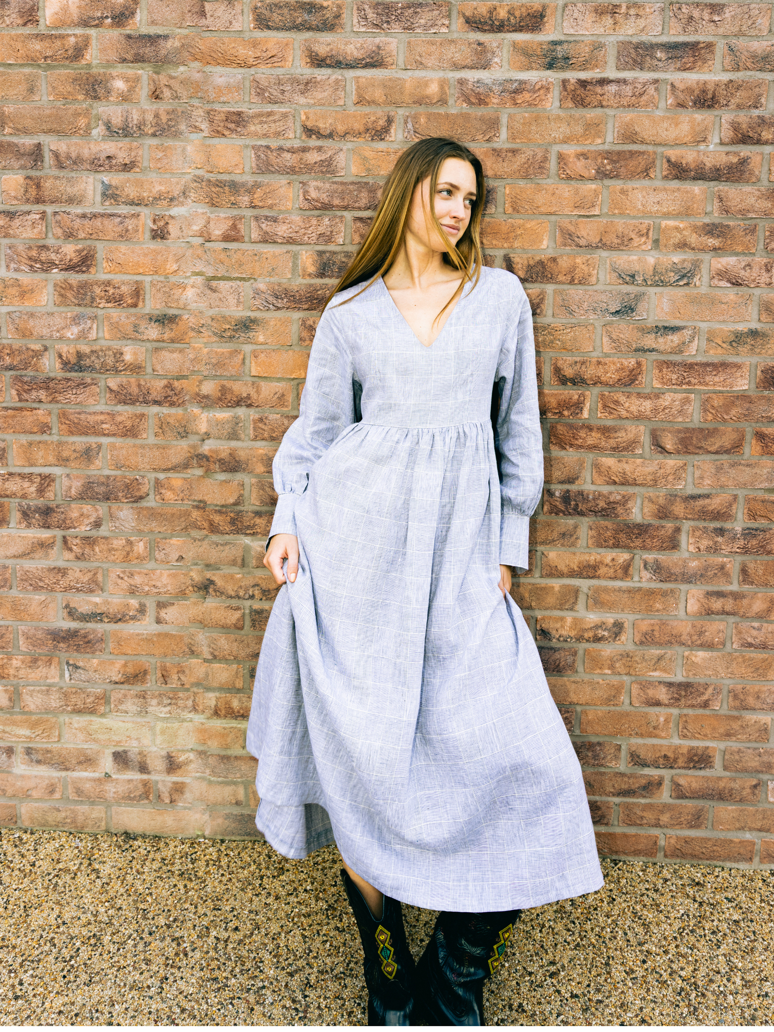 Woman in a long light grey dress standing against a brick wall.