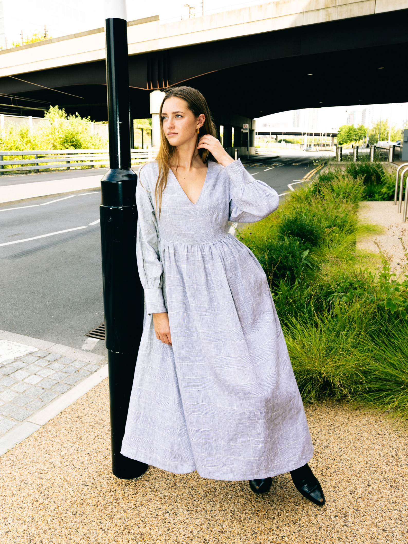 Woman in a long light gray dress standing outdoors near a road and greenery.
