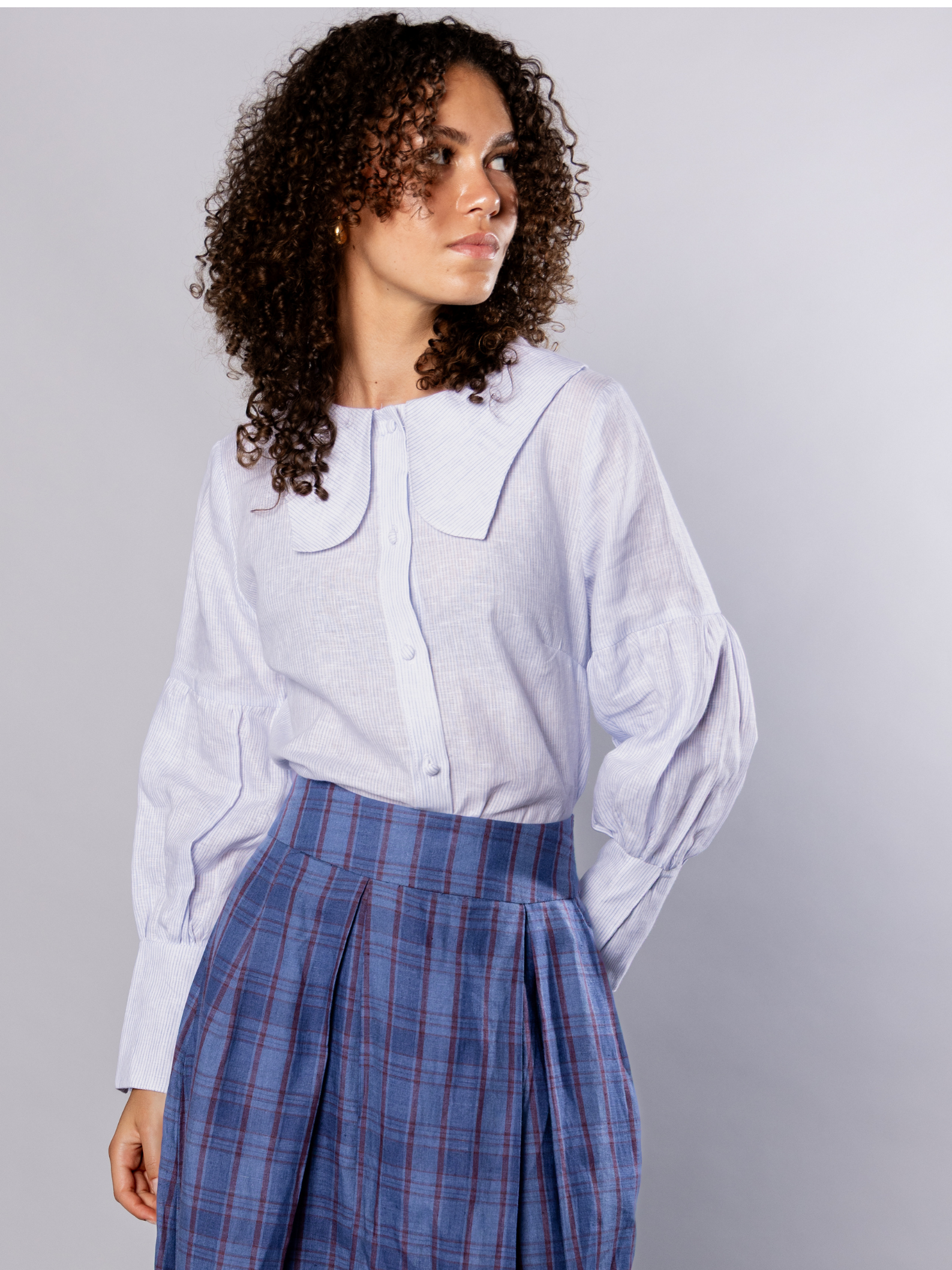 Woman wearing a light blue shirt with a tulip collar and navy blue plaid pants against a grey background