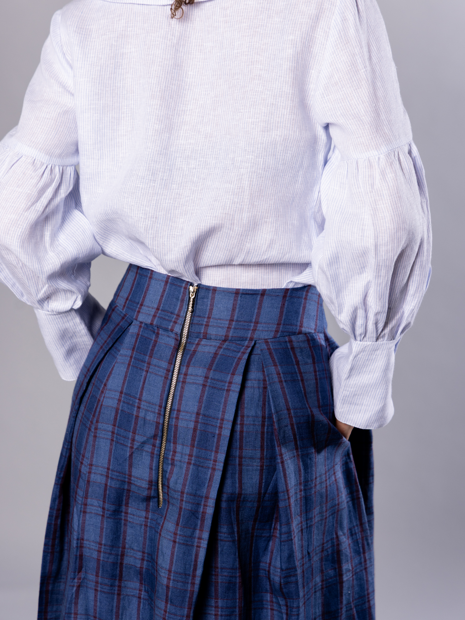 Person wearing a white and light blue blouse with puffed sleeves and navy blue plaid skirt with an exposed zip on a grey background