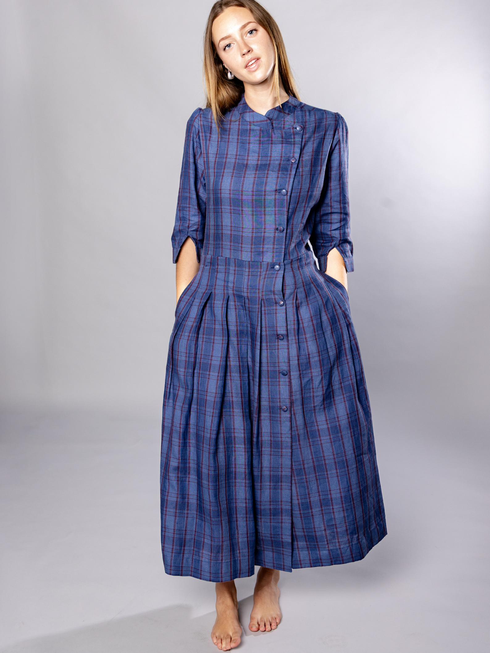 Woman wearing a navy blue plaid dress with side buttons and pockets against a grey background