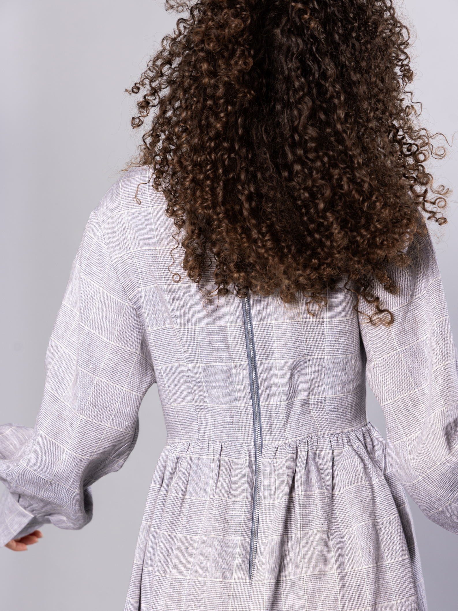 Person wearing a light grey checkered dress with long sleeves and exposed zip on a plain background