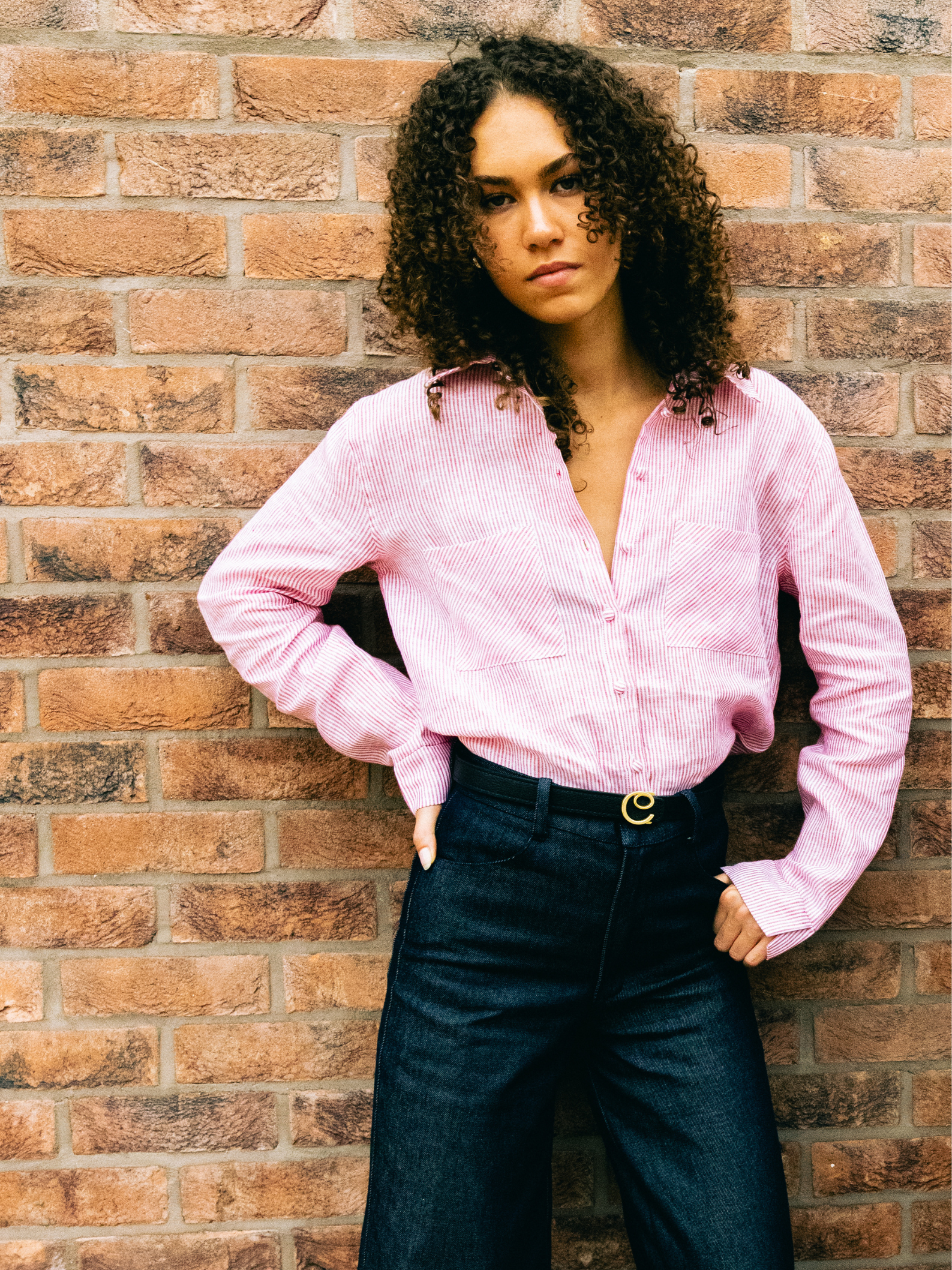 Woman wearing a red stripe shirt and dark navy pants standing against a brick wall