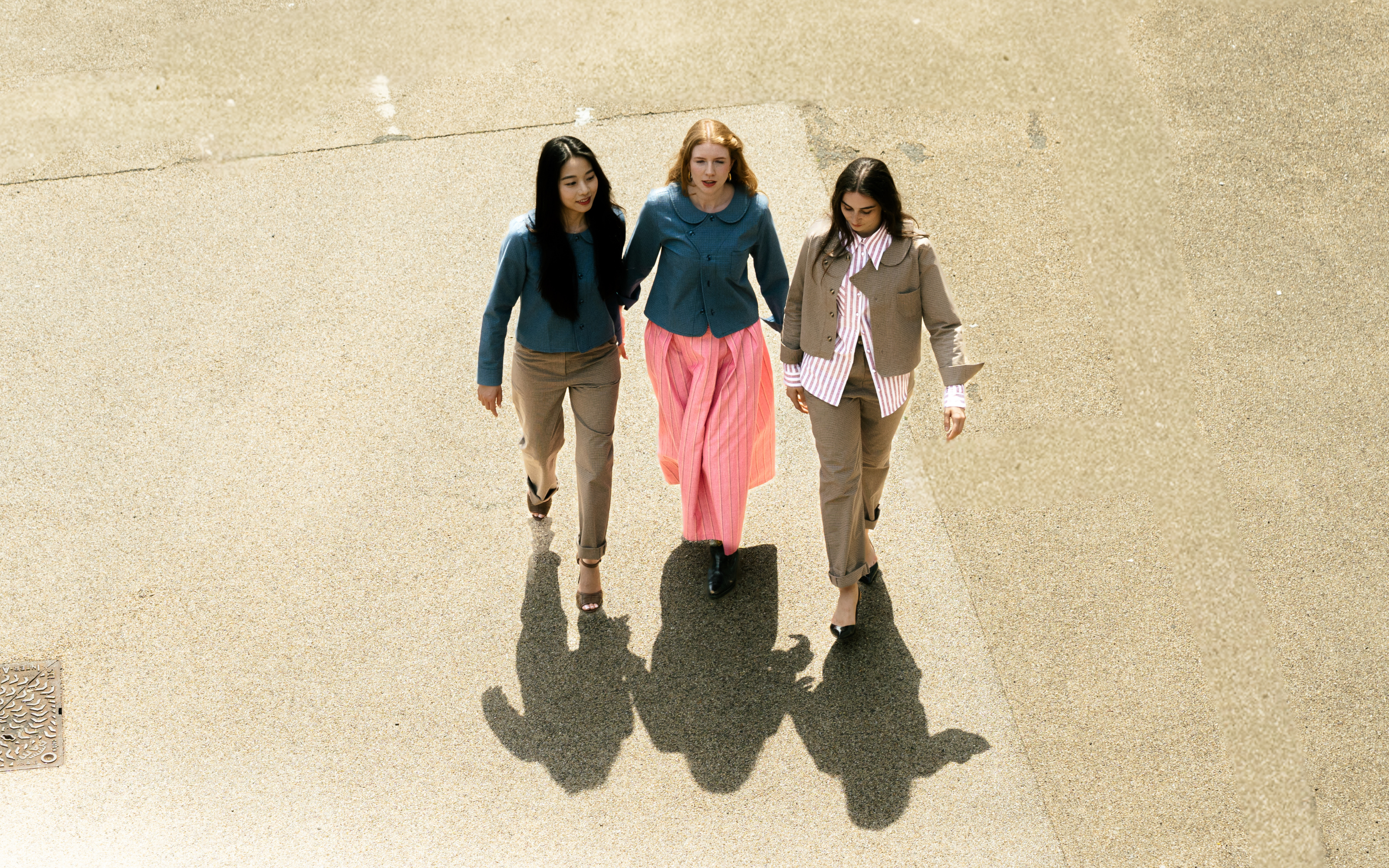Three women walking arm in arm on a concrete path from an overhead angle. The woman on the left wears a teal top and beige pants, the woman in the middle wears a teal top with a pink skirt, and the woman on the right wears a beige blazer over a pink and white striped shirt with beige pants.