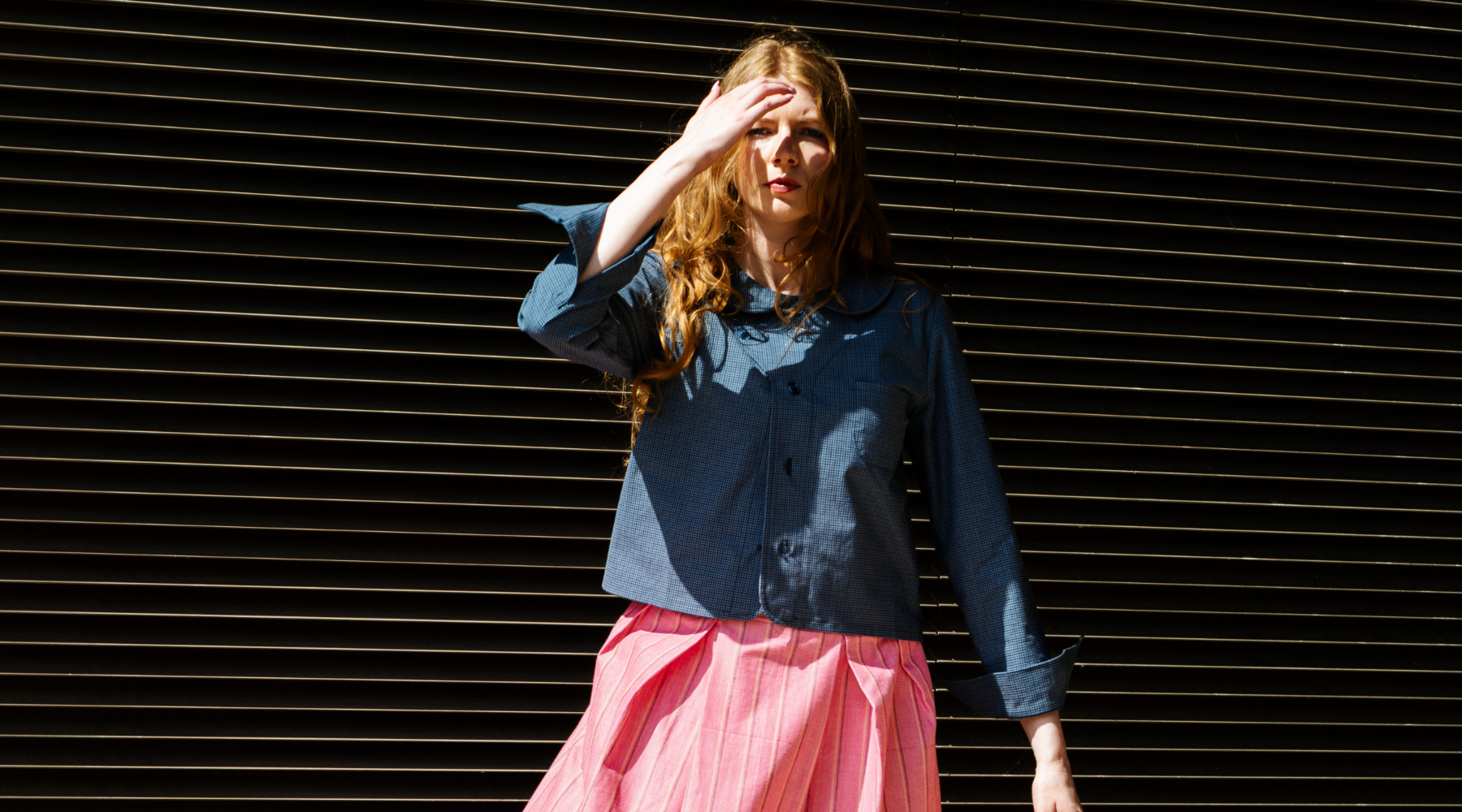 woman with red hair wearing a blue jacket is hiding her face from the sun with her arm against a black background 