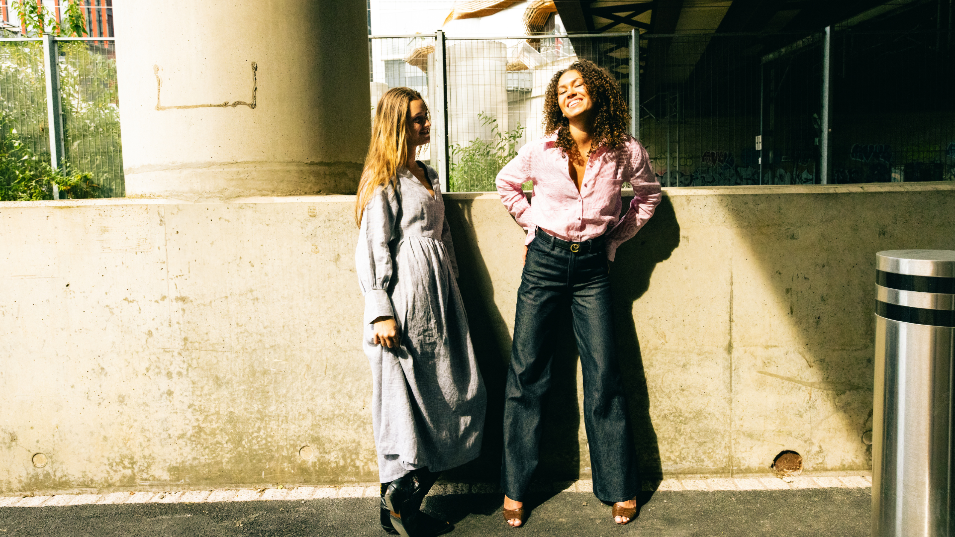 2 women, one blonde, one with black curly hair are talking and smiling under a railway bridge