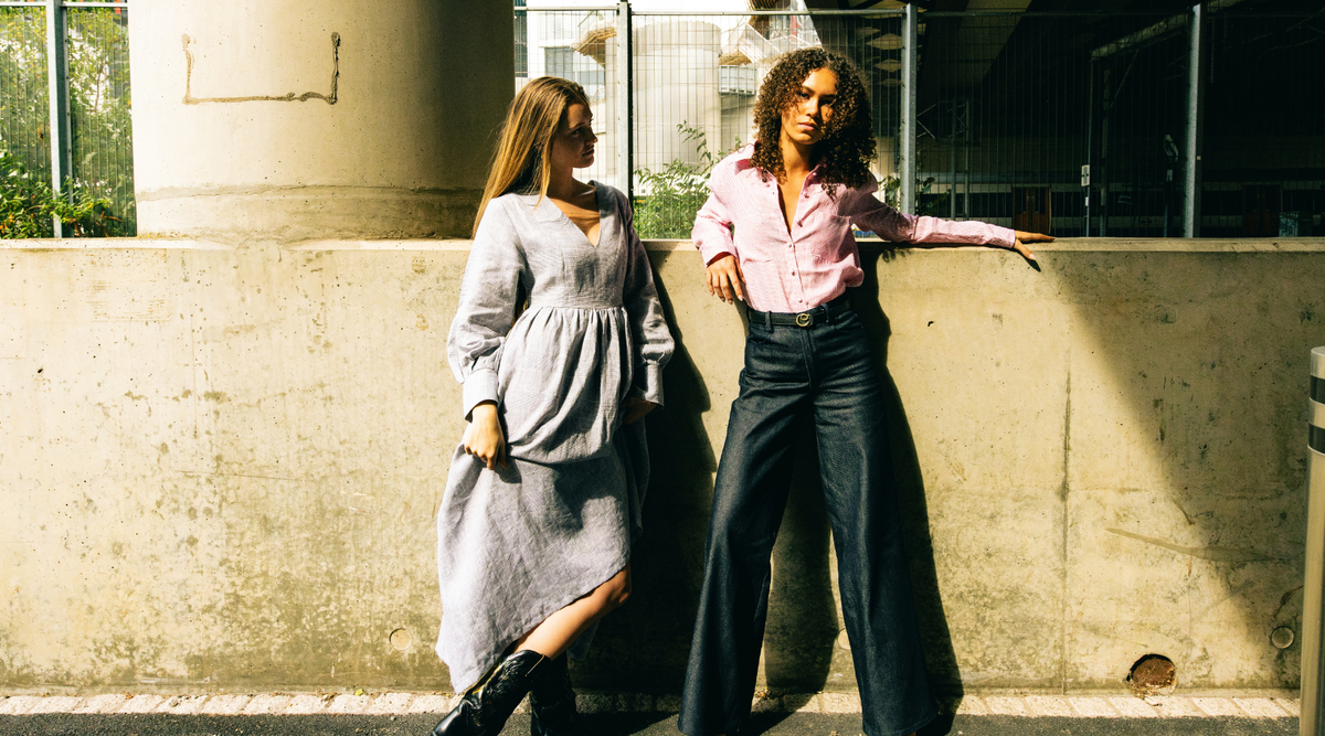 two women leaning against a concrete wall, one wearing a long grey dress, the other wearing navy jeans and a pink shirt