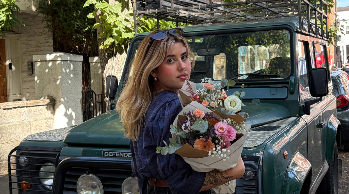 a young woman holding bunch of flowers standing in front of a green jeep car with sunglasses in her hair