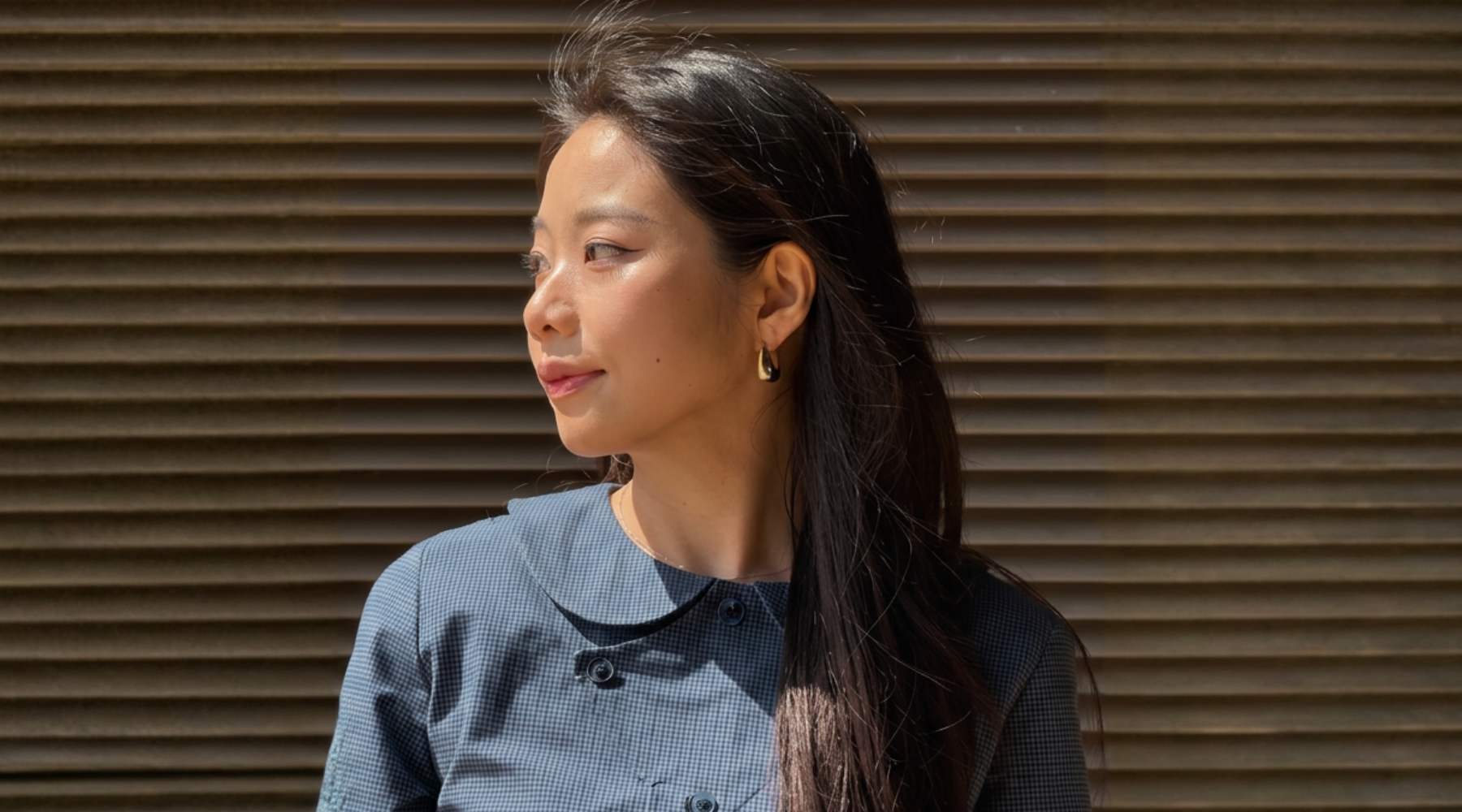 asian woman looking to the distance wearing gold earrings and a blue jacket against a grey background