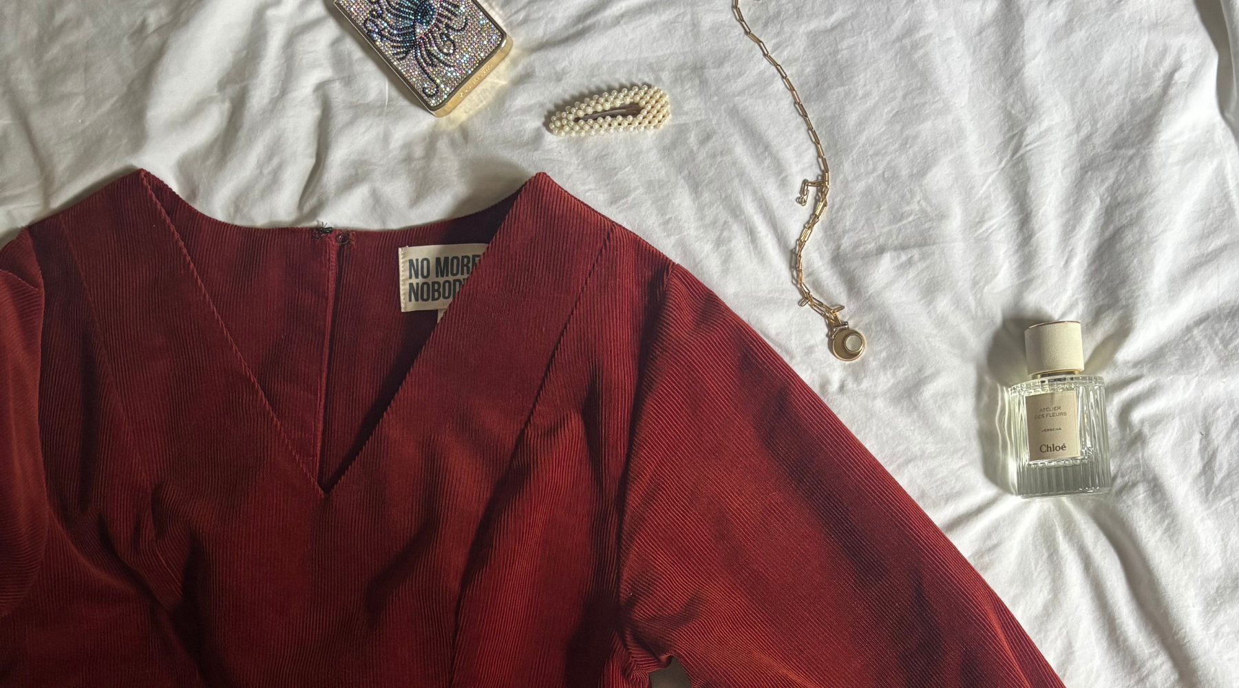 red dress on a white bedding with scattered nude items, perfume, necklace, hair clip and a phone