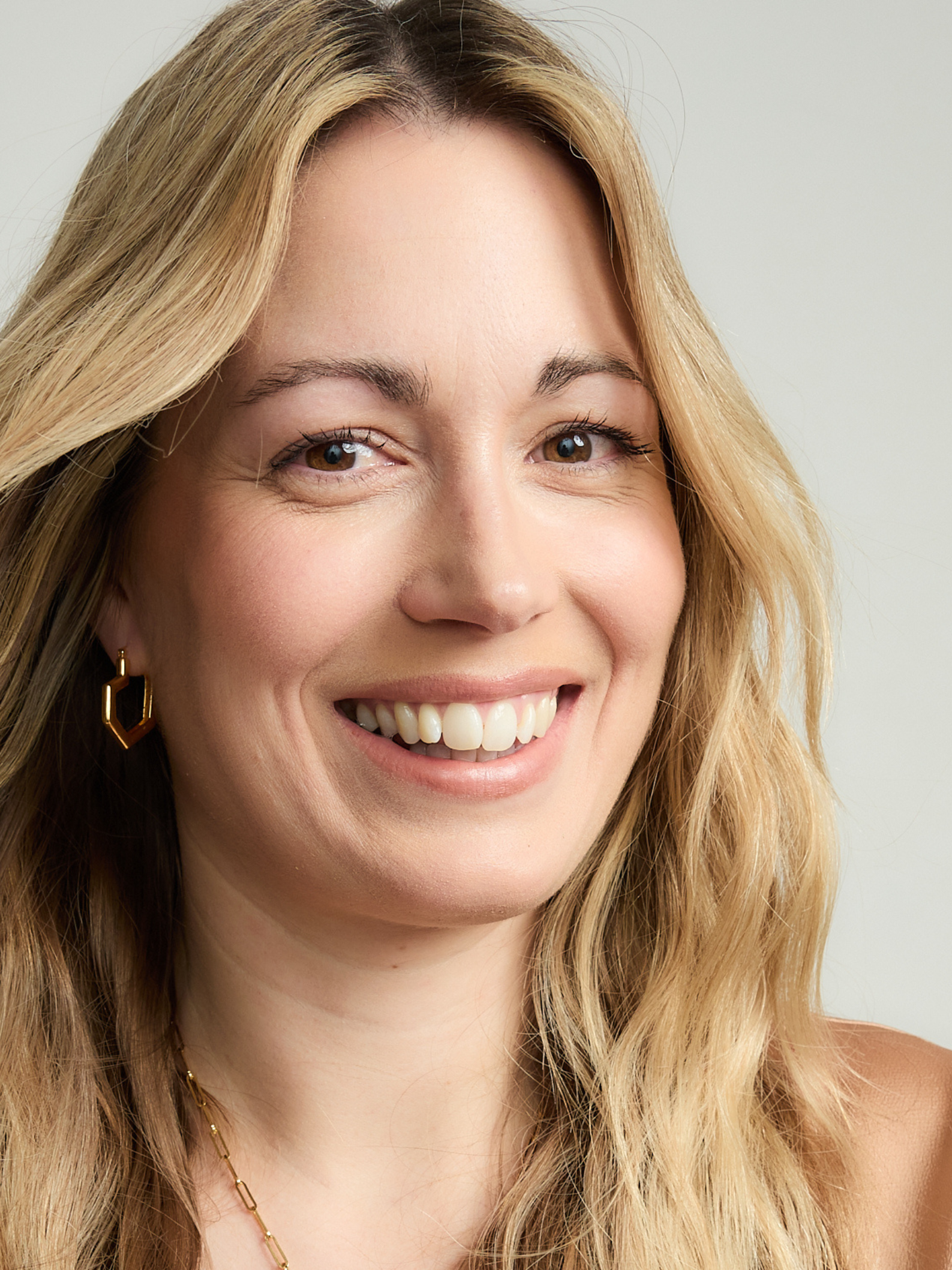 Woman with blonde hair and gold heart hoop earrings smiling against a neutral background