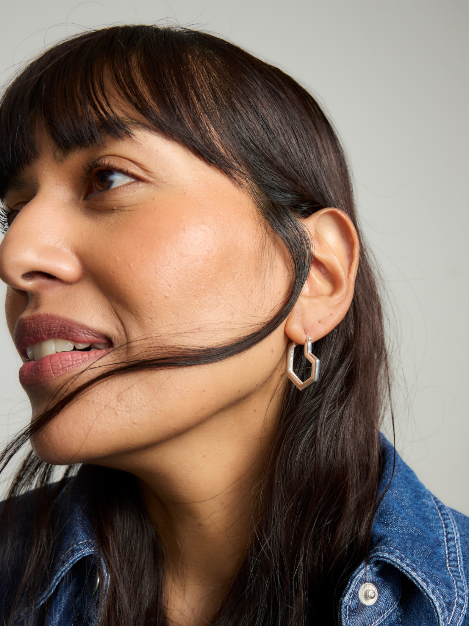 Woman with a strand of hair over her face, wearing a denim jacket and silver heart hoop earring.