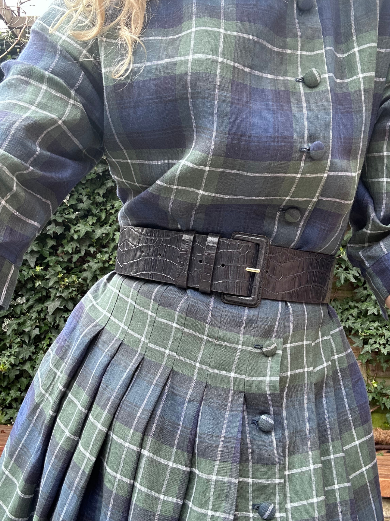 Person wearing a plaid outfit with a belt in front of a green bush