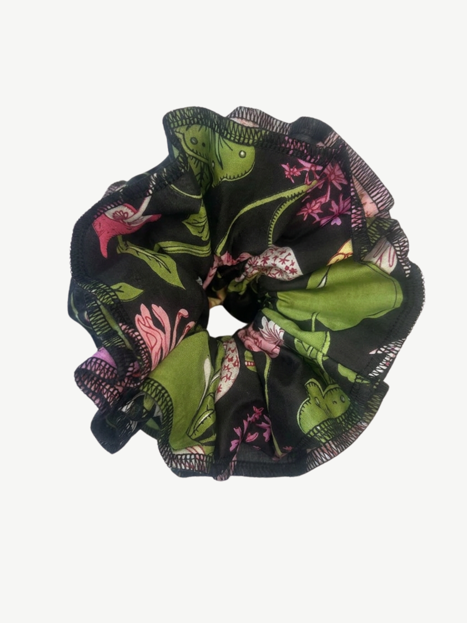 A black fabric hair scrunchie featuring a botanical print with green leaves, pink flowers, and small yellow accents, shown against a plain white background.