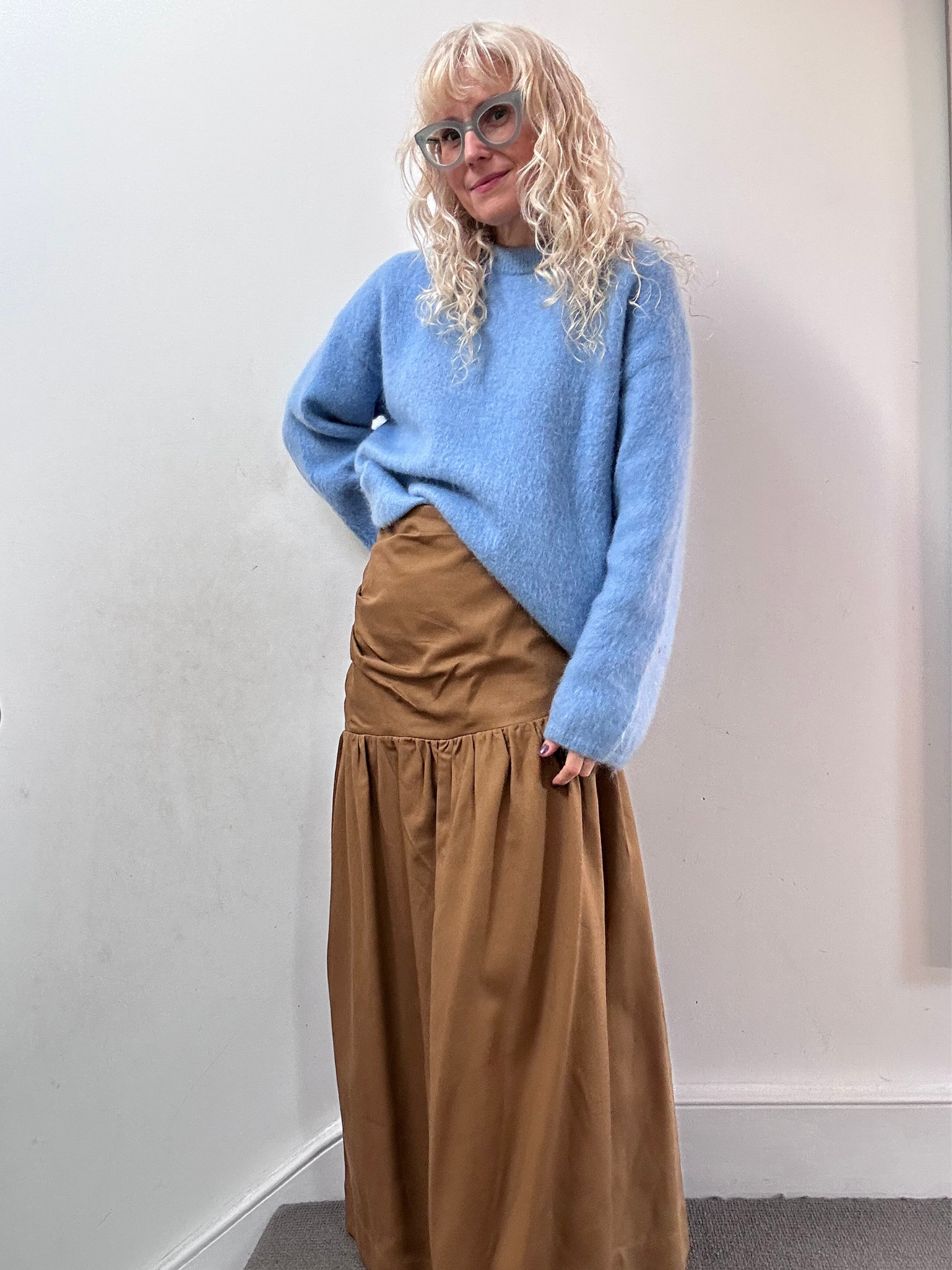 Person wearing a blue sweater and brown skirt against a white wall