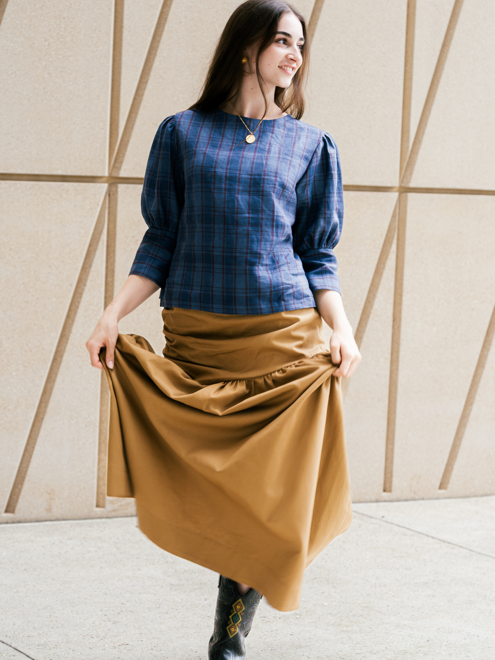 Woman wearing a blue plaid shirt and brown skirt against a geometric wall.