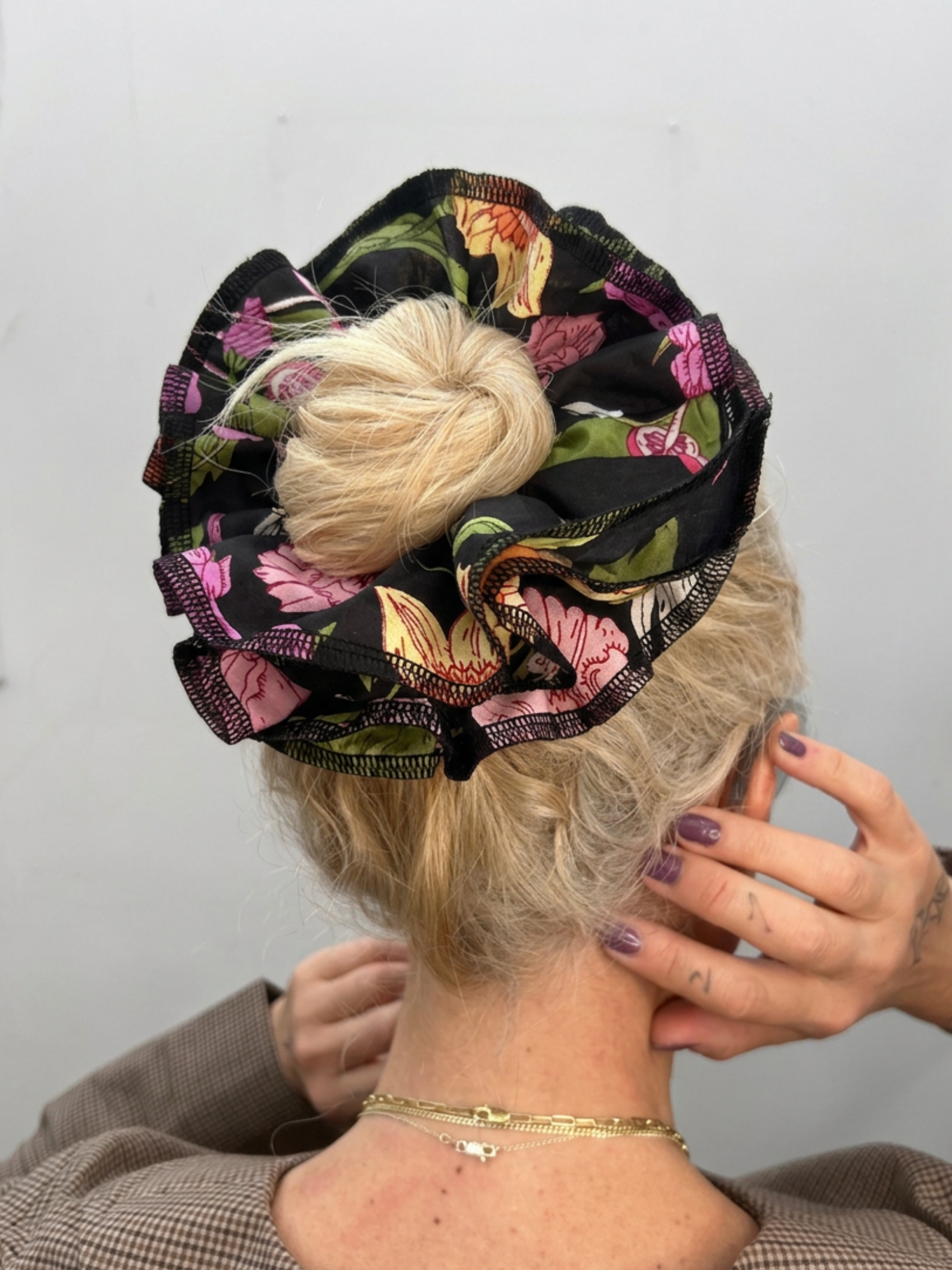 A blonde person shown from the back wearing a large floral hair scrunchie in black with pink, green, and yellow floral print. Their hands are visible behind their head, and they are wearing a gold chain necklace against a plain light gray wall.