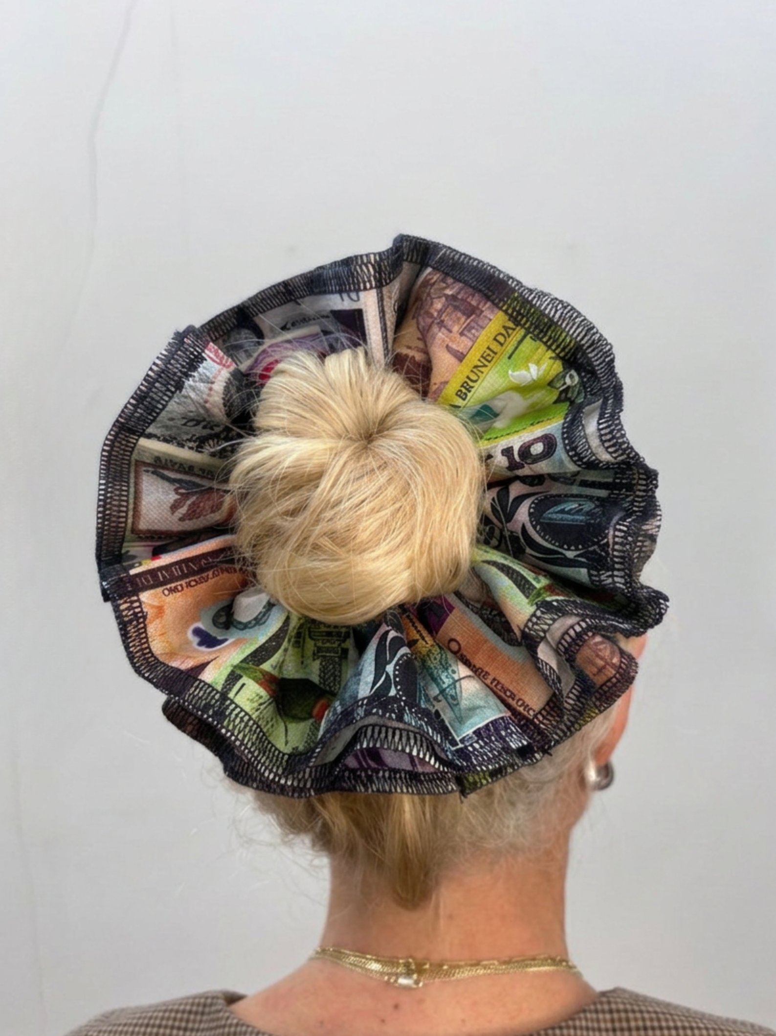 A blonde woman viewed from the back wearing a large, ruffled hair accessory made from printed banknotes, with a blonde bun in the center.