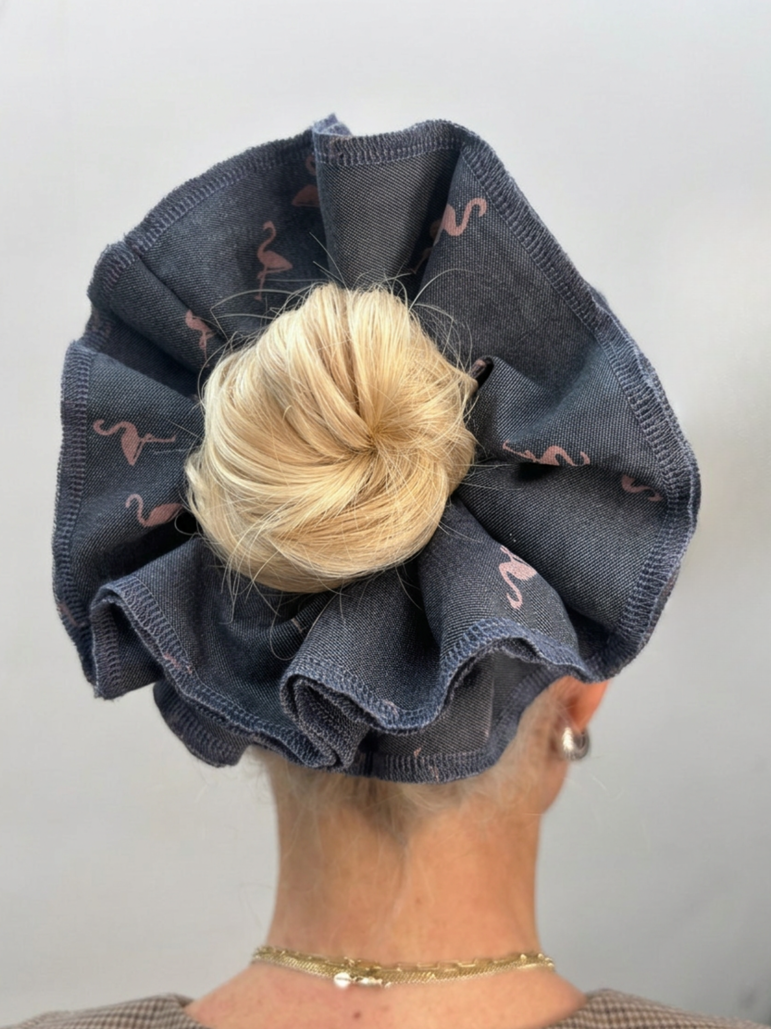 A close-up back view of a person wearing a large fabric scrunchie on their hair. The scrunchie is made of dark gray denim-like fabric with a subtle pink flamingo print, and the person's blonde hair is tucked inside.