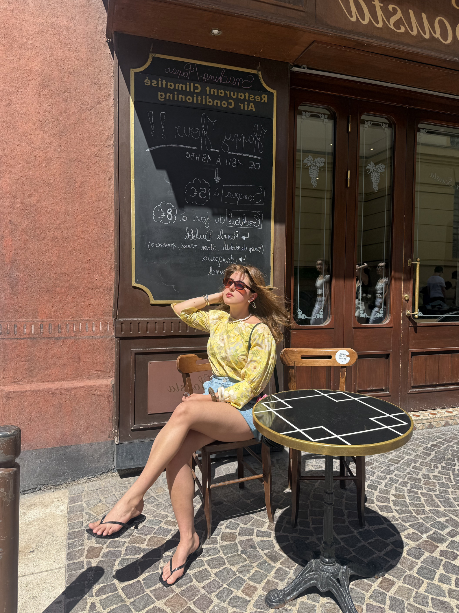 Woman in yellow top and denim shorts sitting at a table outside a café.