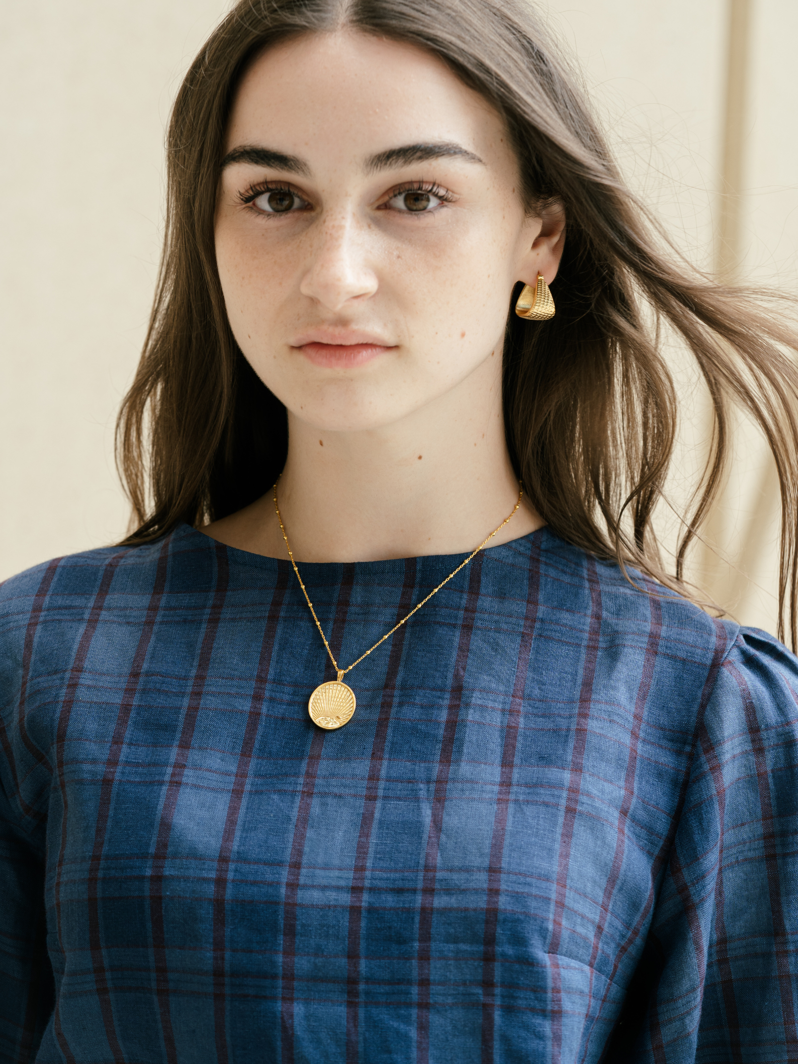 Woman wearing a blue plaid top with gold jewelry against a neutral background