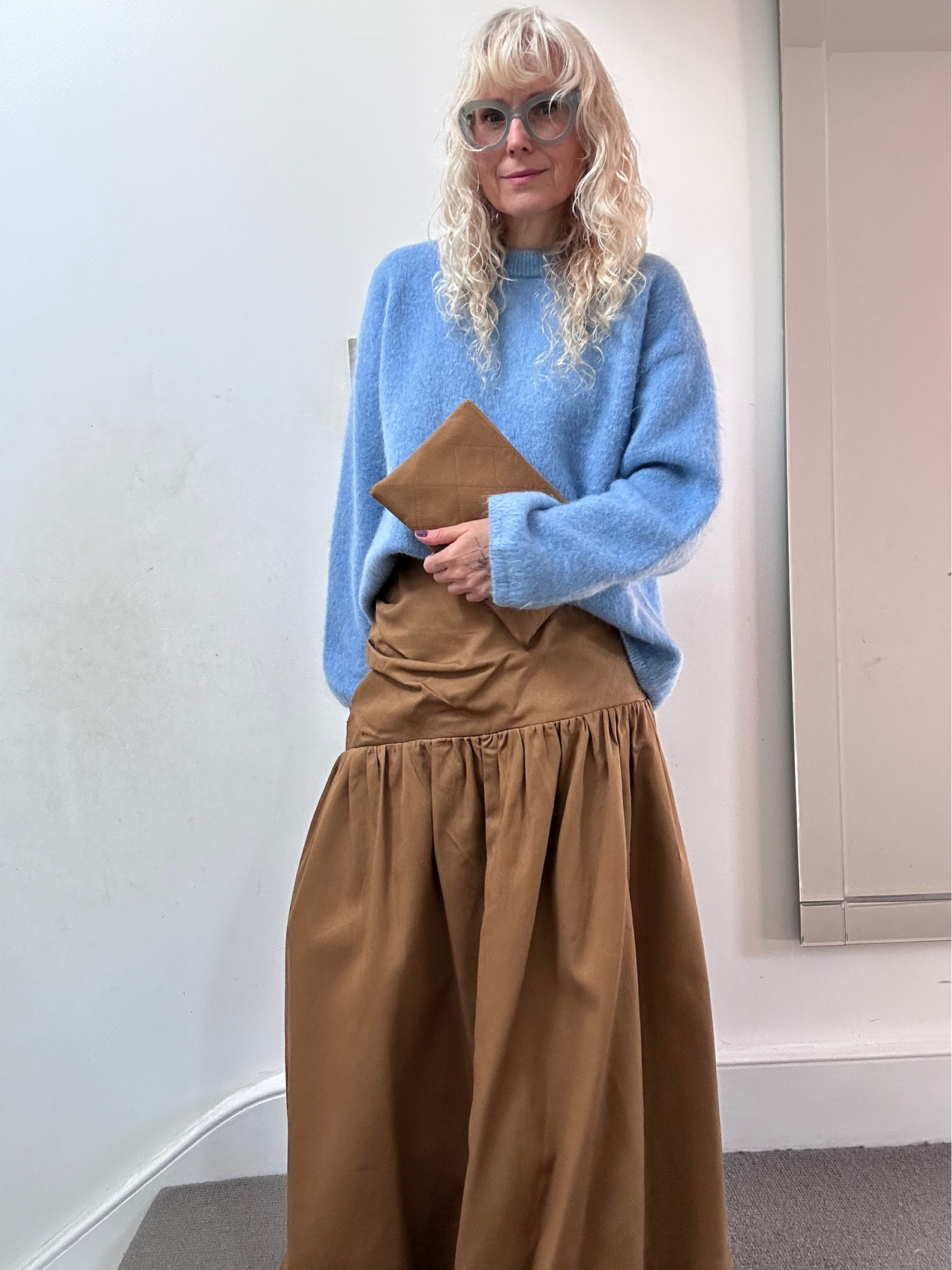 Person wearing a blue sweater and brown skirt holding a brown bag indoors.