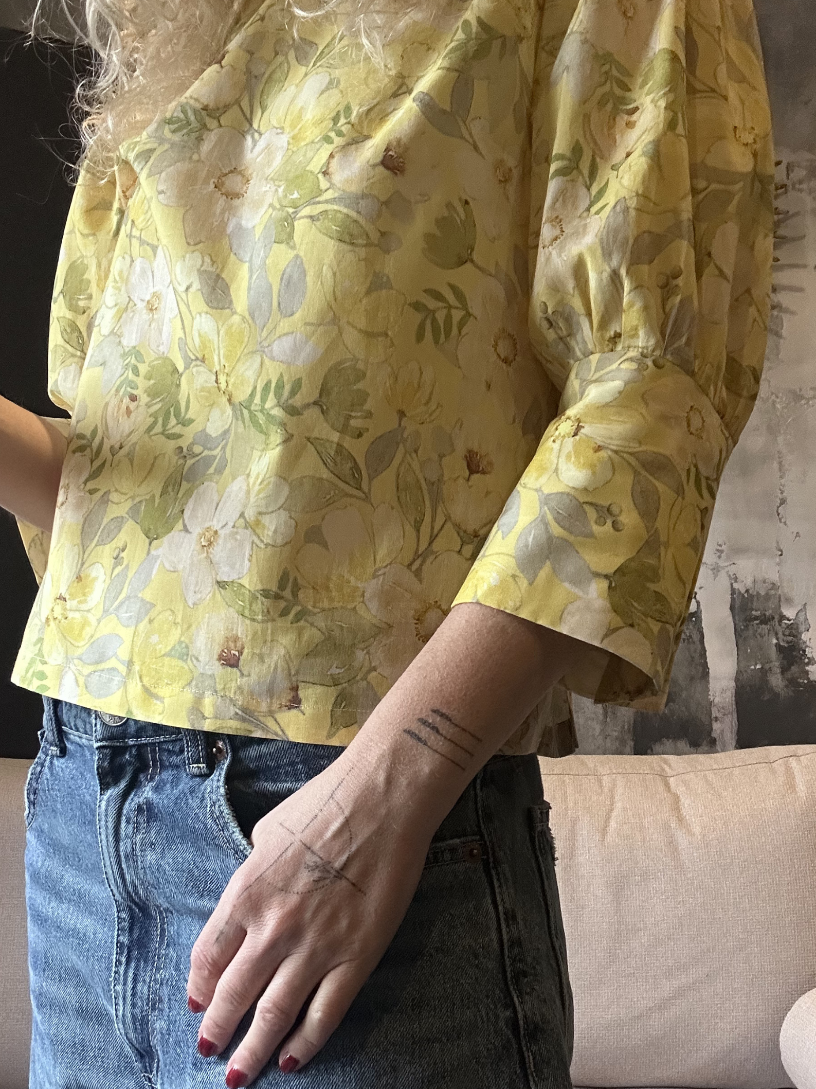 Person wearing a yellow floral blouse with rolled-up sleeves, standing indoors.