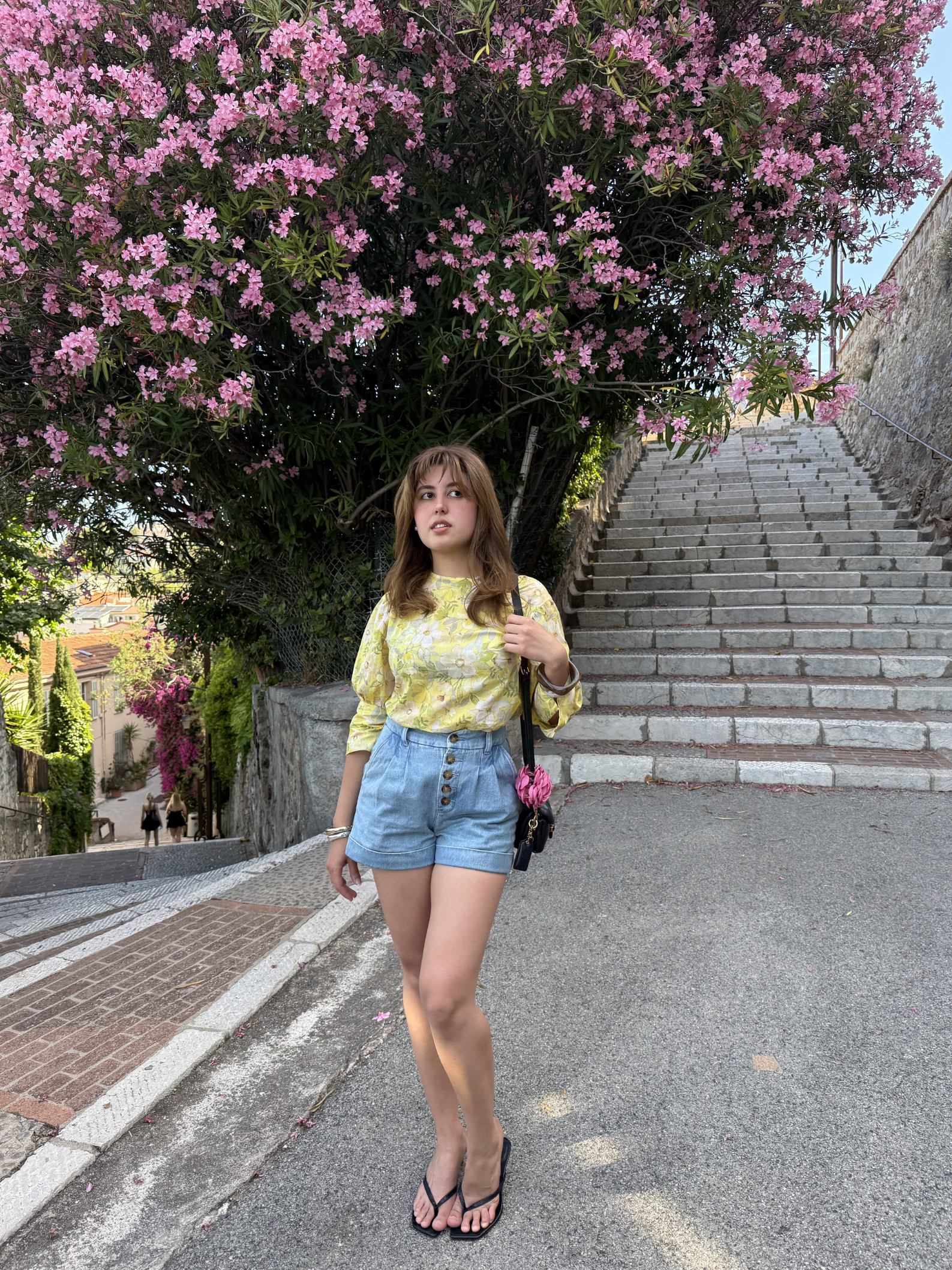 Woman in yellow top and denim shorts standing in front of a flowering tree on a street.