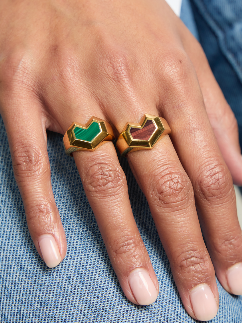 Close-up of a hand wearing two heart rings with green and red stones on a blue fabric background